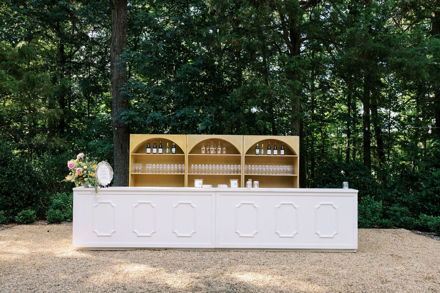 Elegant outdoor bar setup with white paneled counter and gold arched shelving unit displaying glassware amid lush trees.