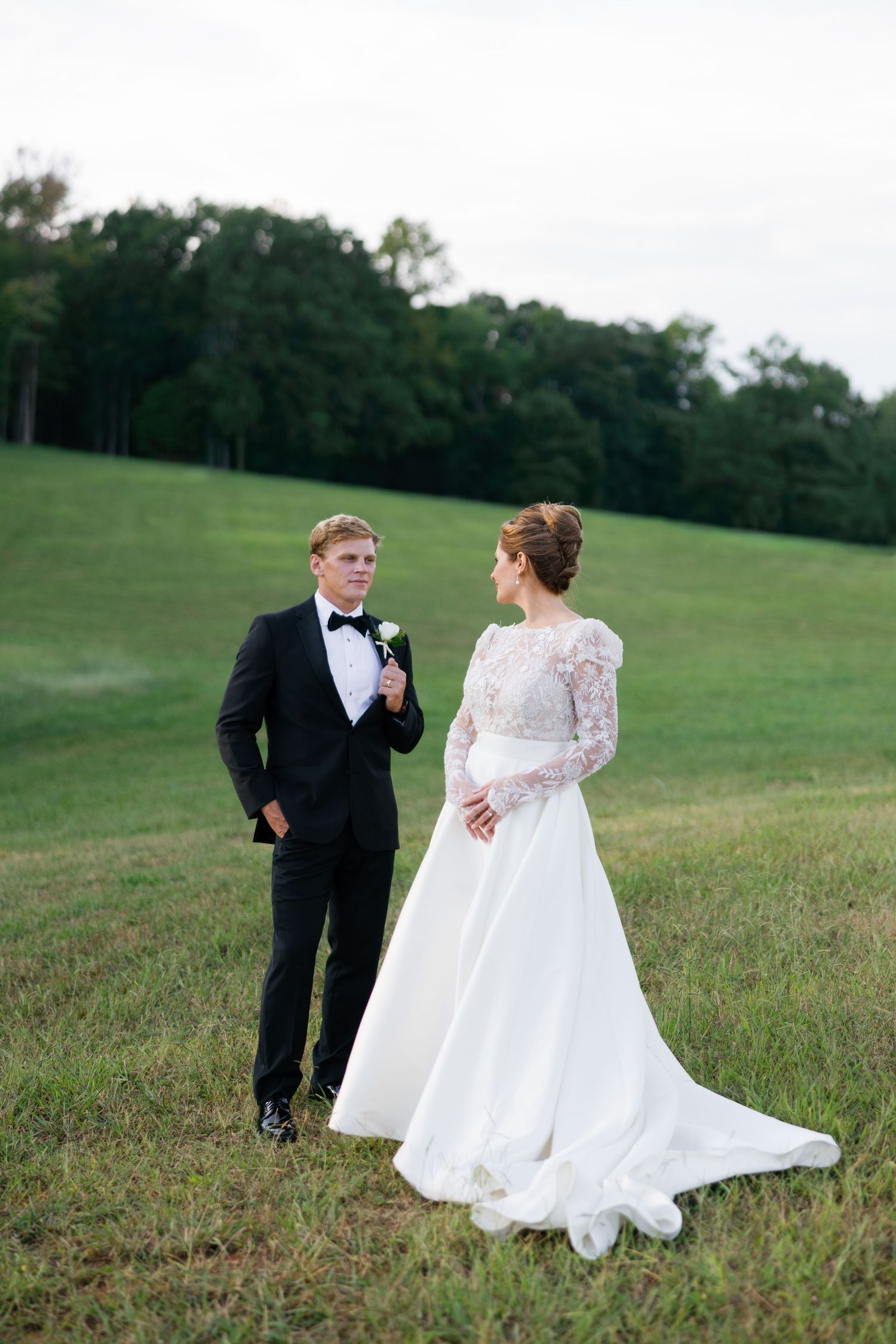 A bride in a white lace gown and groom in black tuxedo stand together on a green grassy field at dusk.