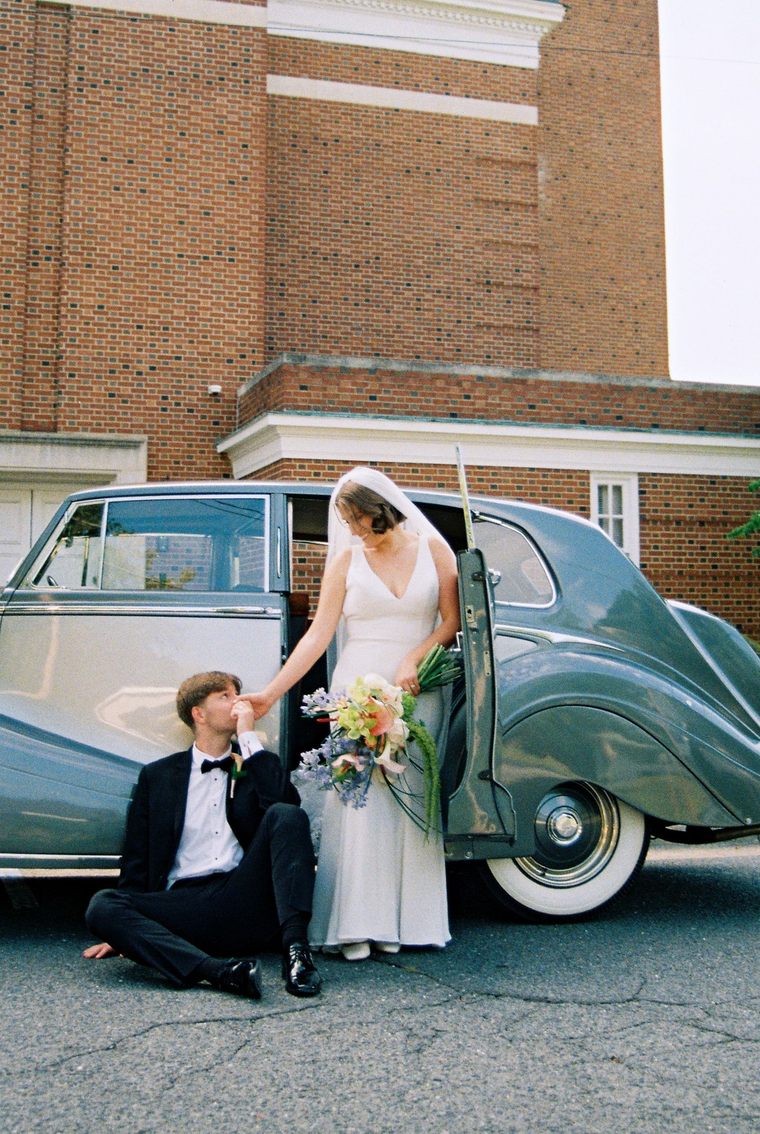 Elegant vintage car serves as backdrop for romantic wedding photo outside brick church building.