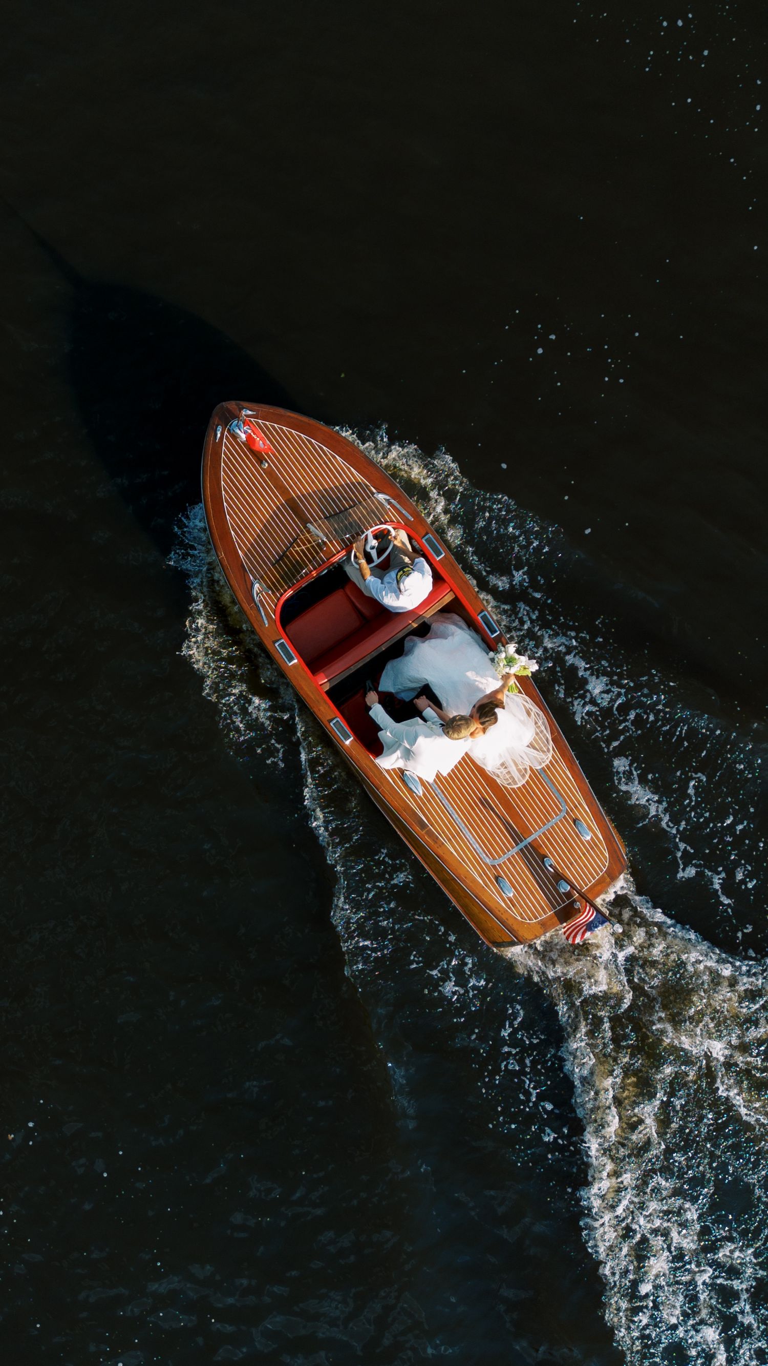 Aerial view of a classic wooden speedboat cutting through dark water creating a dramatic wake pattern.