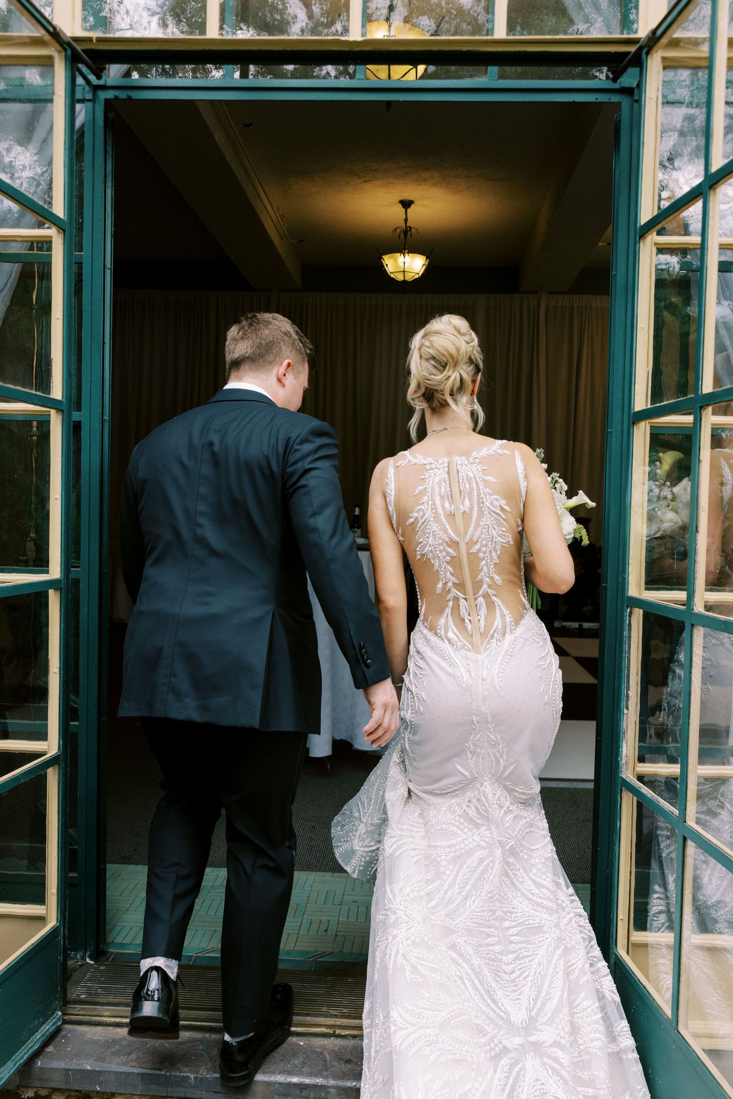 Bride and groom holding hands entering elegant venue, bride wearing stunning backless lace mermaid wedding gown with intricate detail.