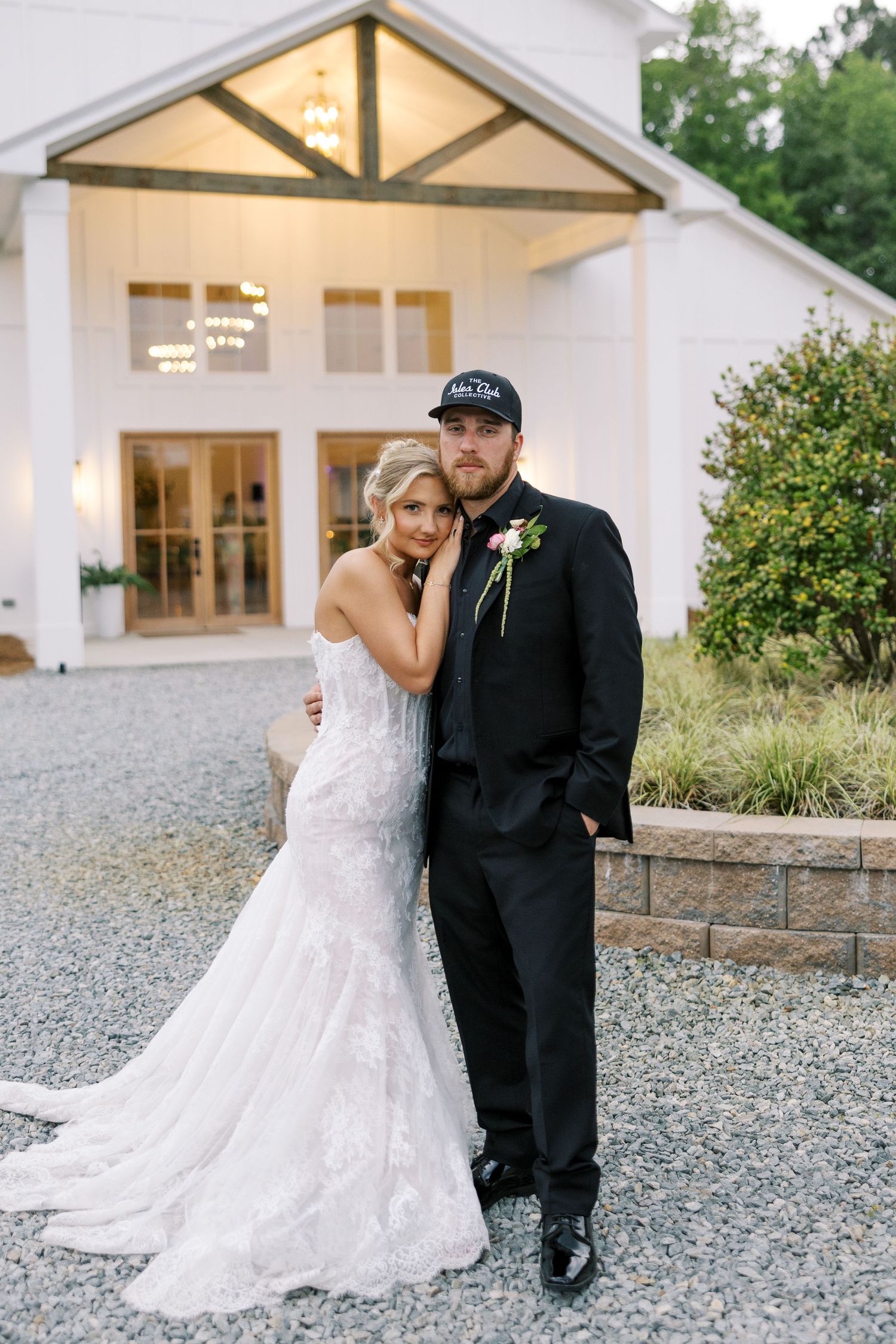 Bride in lace mermaid gown leans on groom in black suit and cap outside white wedding venue at dusk.