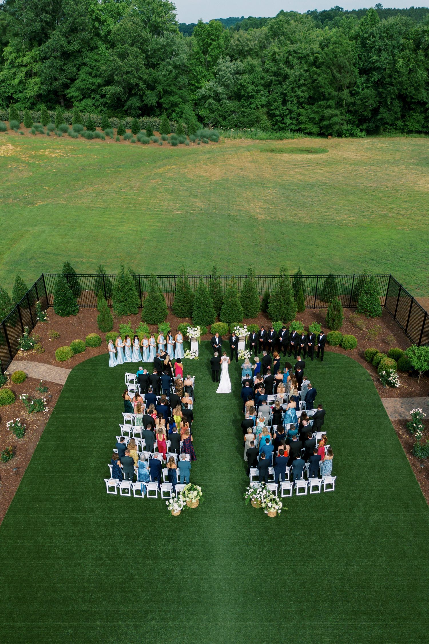 Aerial view of an elegant outdoor wedding ceremony on lush green grass with white chairs and bridal party.