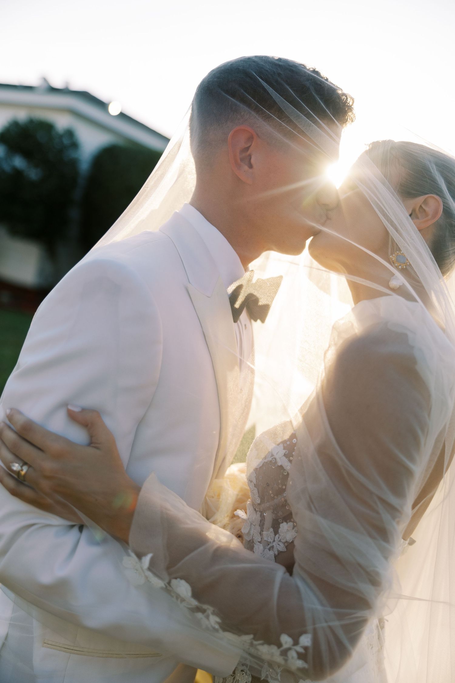 Bride and groom kissing under veil with sunlight glowing behind them.