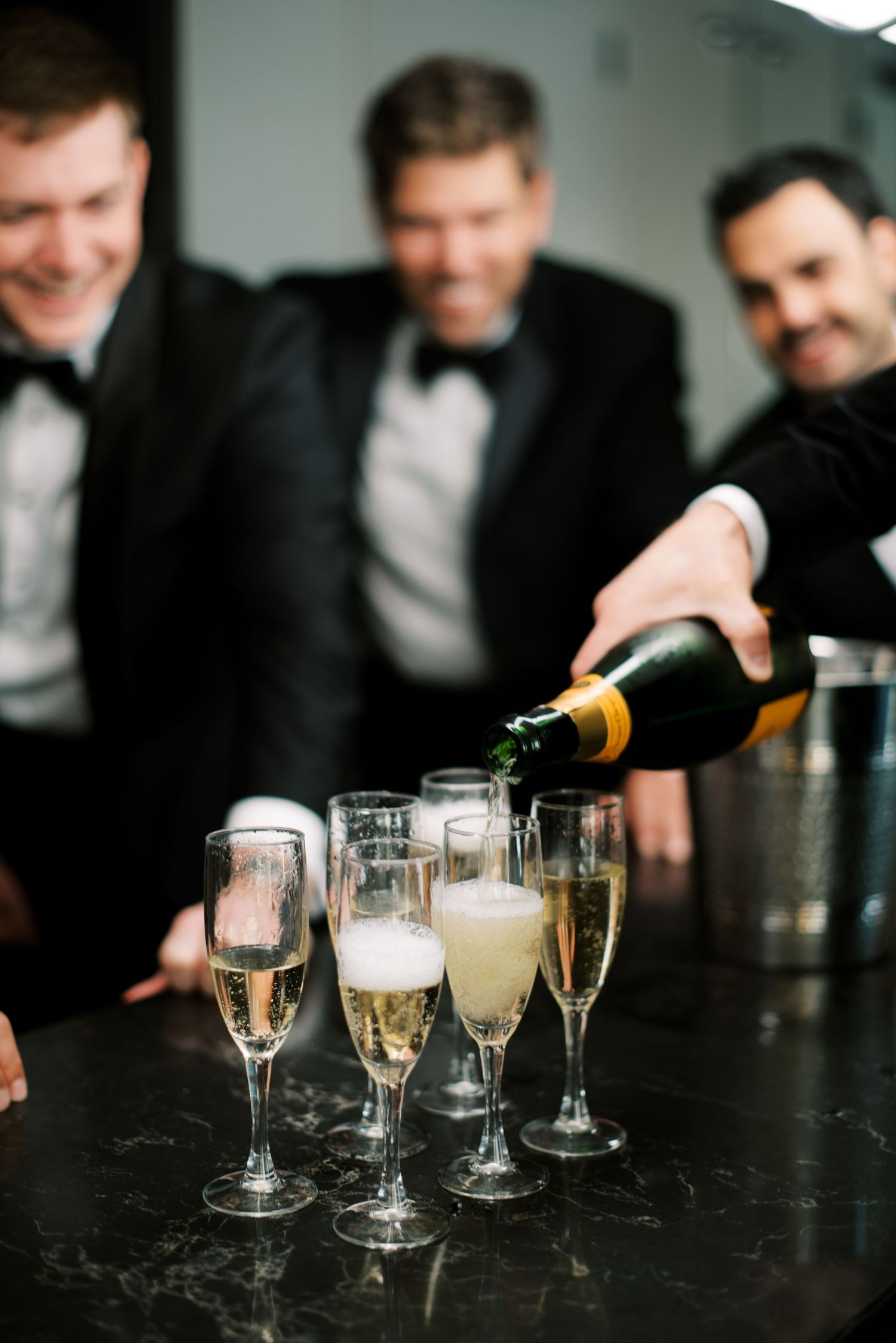 Champagne glasses being filled with bubbly drinks on a dark table while people gather around to watch.