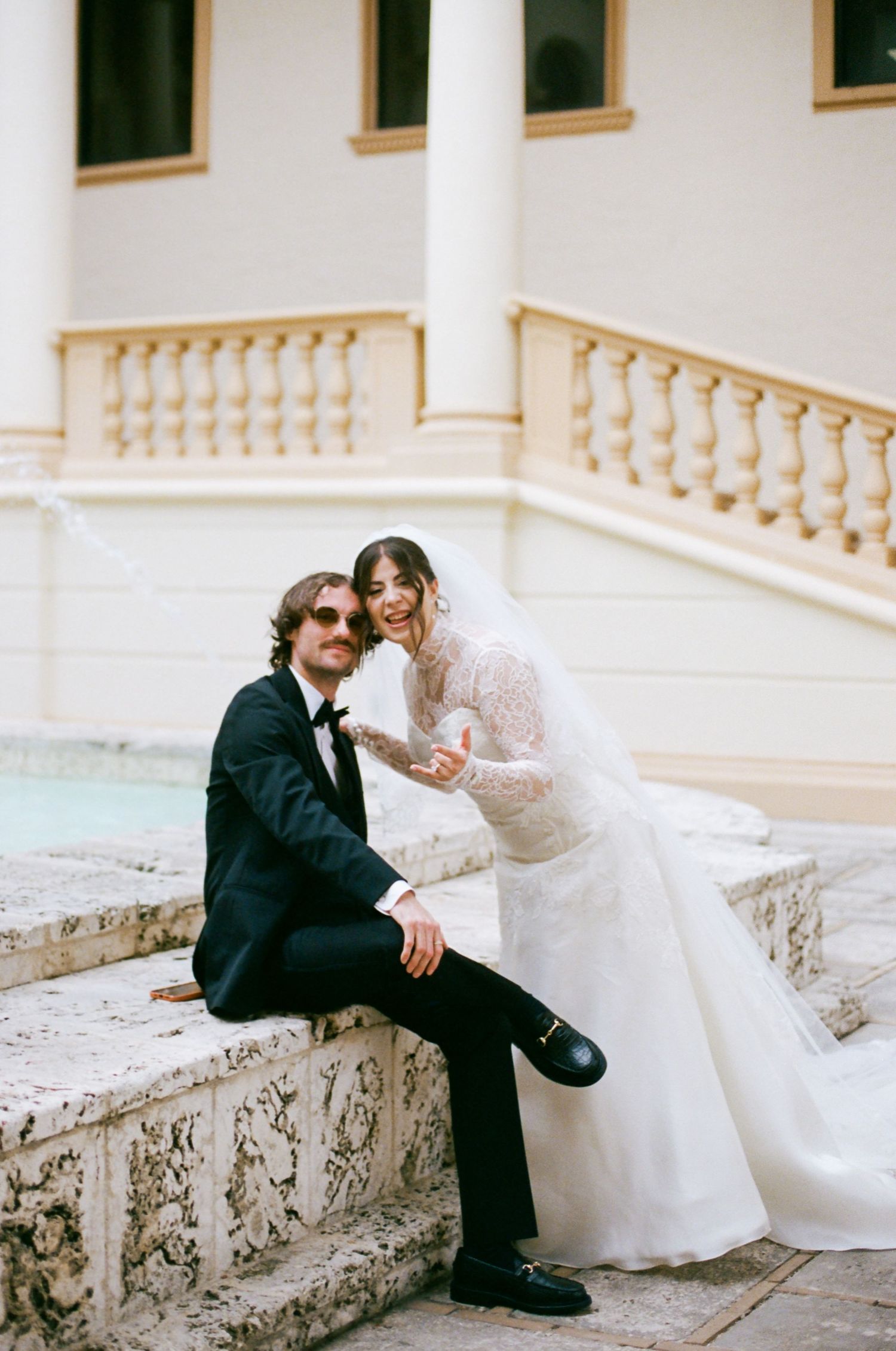 A romantic wedding couple sits together on ornate stone steps of a grand building with white columns and balustrades.
