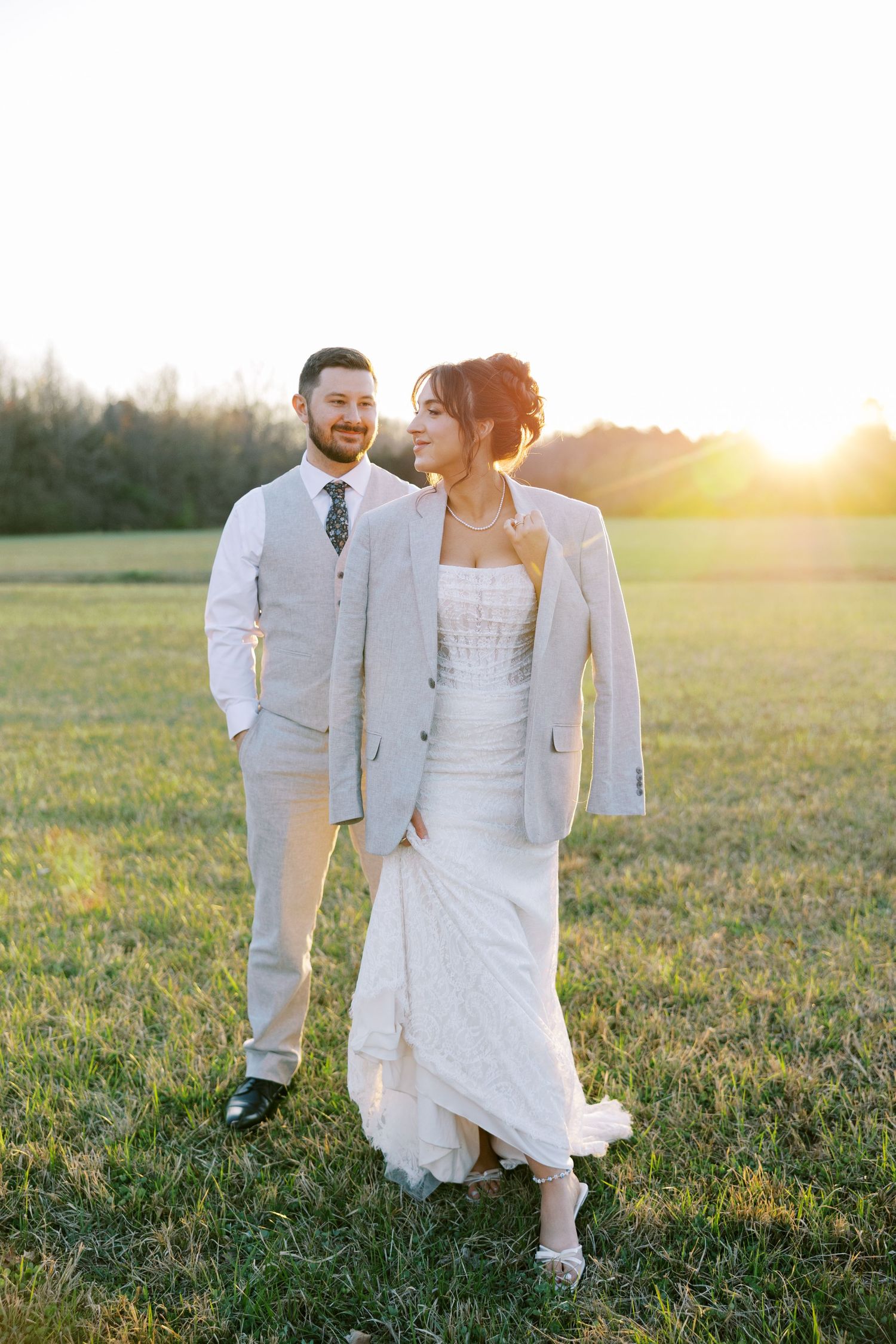 A couple in white wedding attire walks through a sunlit meadow during golden hour.