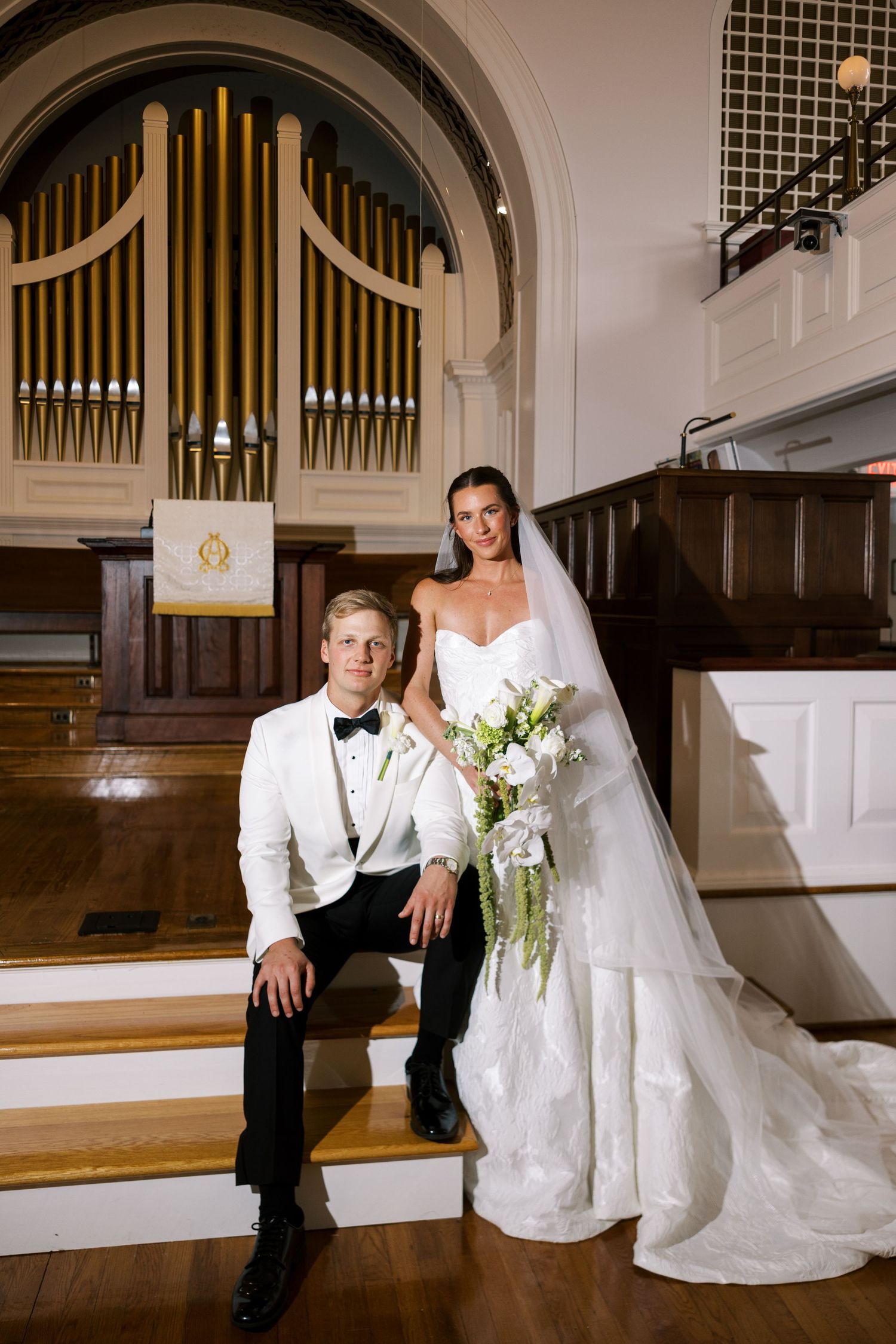 Bride and groom pose on church steps before a grand pipe organ, she holds white florals in a strapless gown with veil.