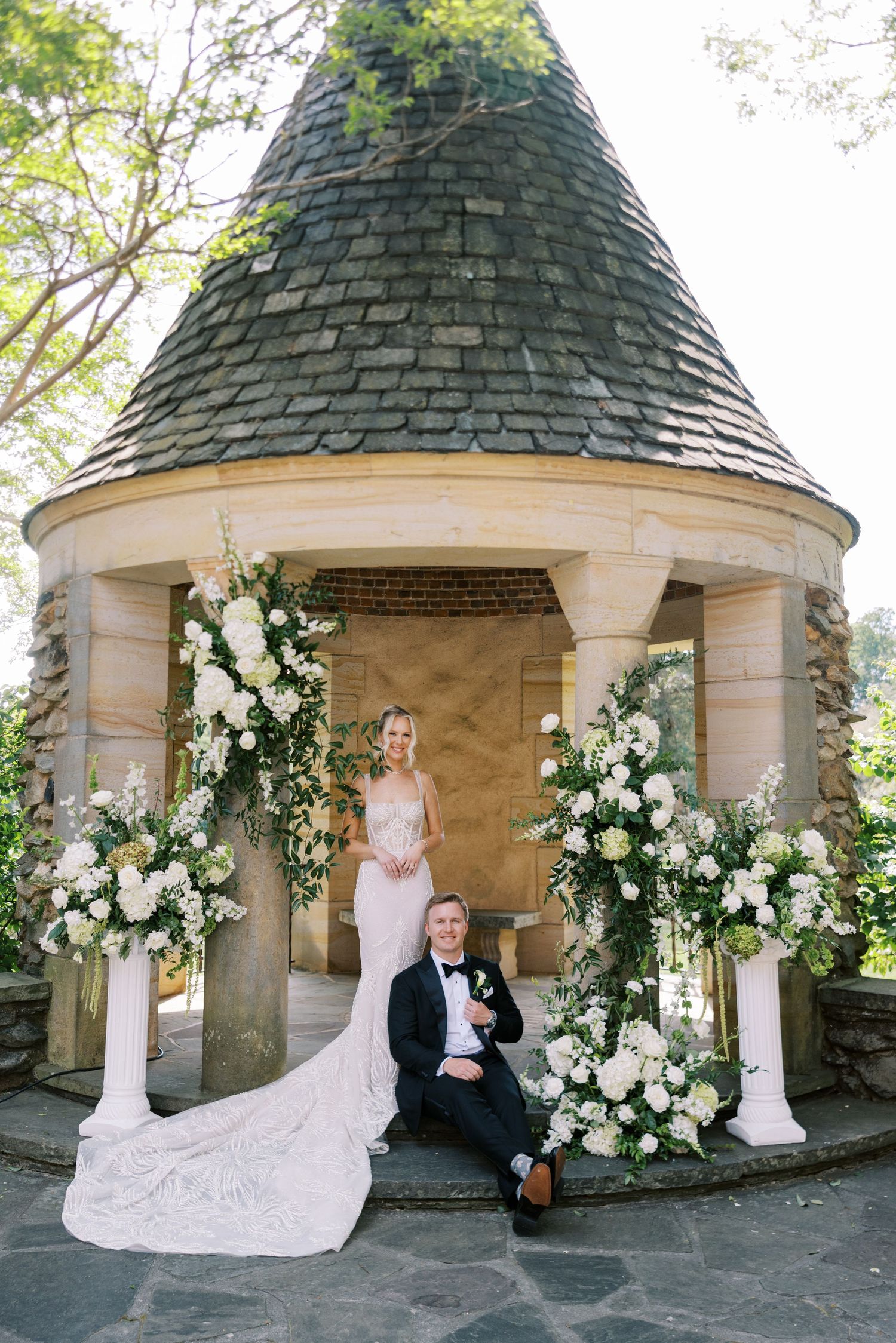 Bride and groom pose at elegant stone gazebo adorned with white floral arrangements for their wedding portrait.
