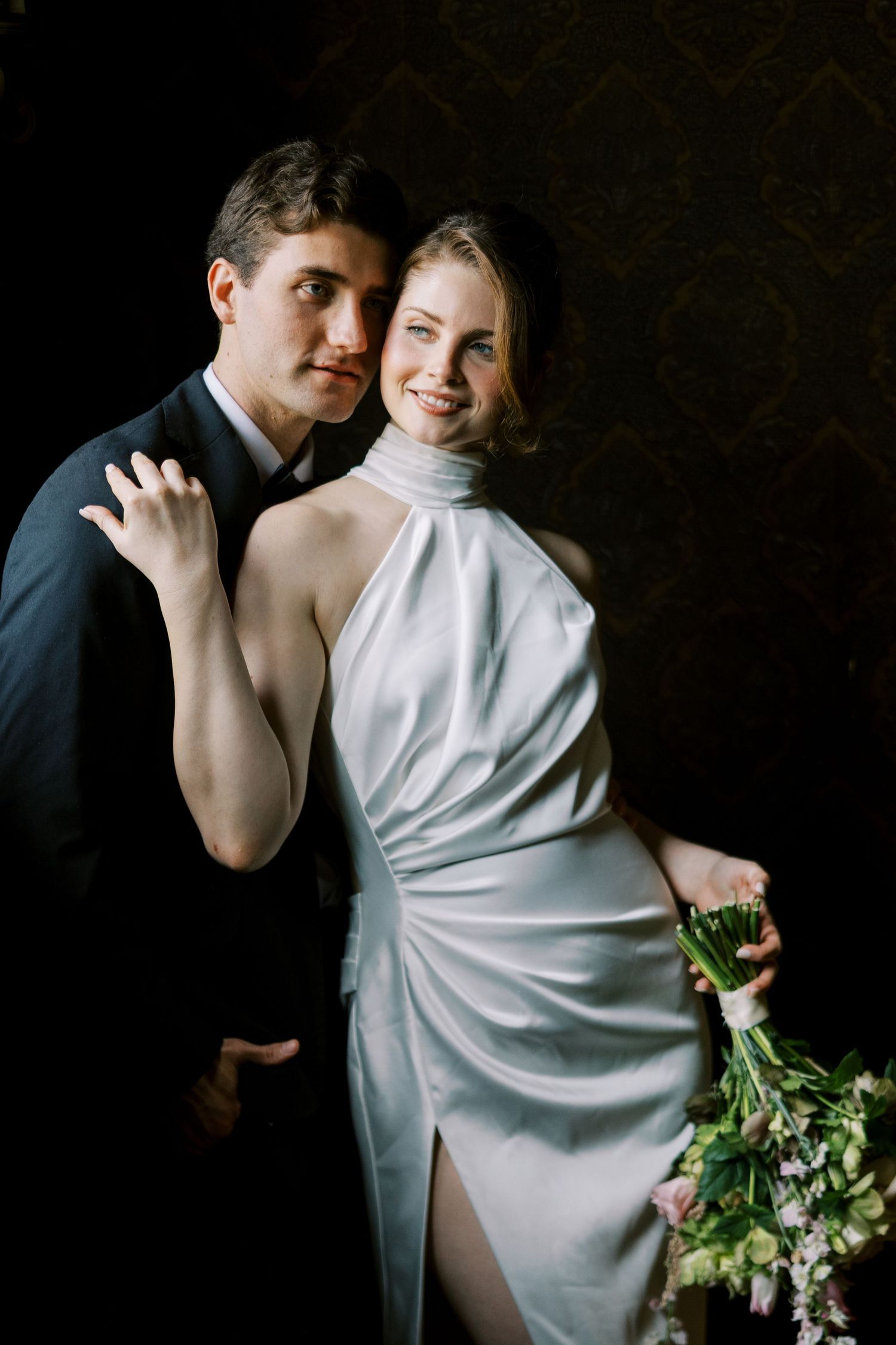 Elegant bride in white halter gown holds bouquet beside groom in black suit against dark background.