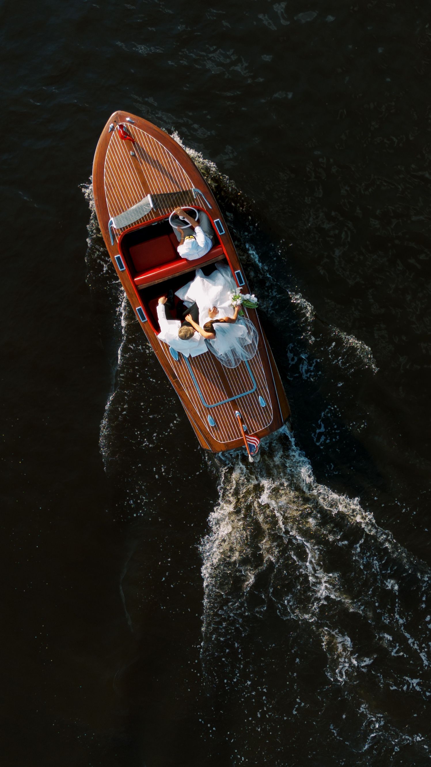 Aerial view of a classic wooden speedboat with a wedding couple in white attire gliding across dark water.