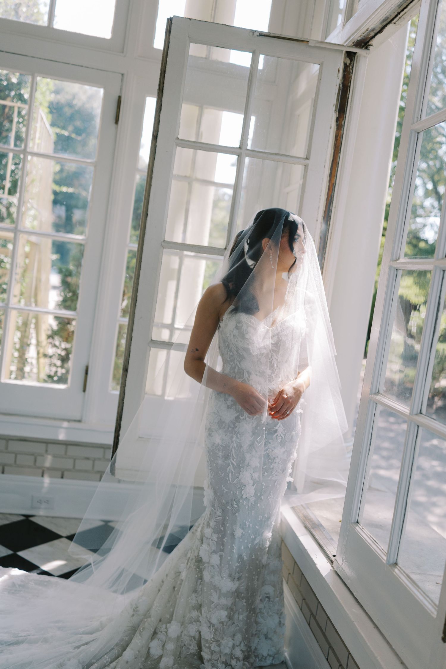 A bride in an elegant lace wedding gown and flowing veil stands by sunlit French doors in a bright interior space.
