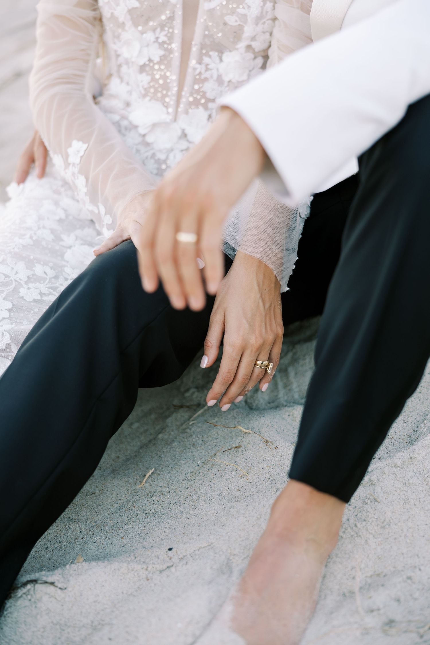 Bride and groom sitting on sand, displaying gold wedding rings on their hands, wearing formal wedding attire.