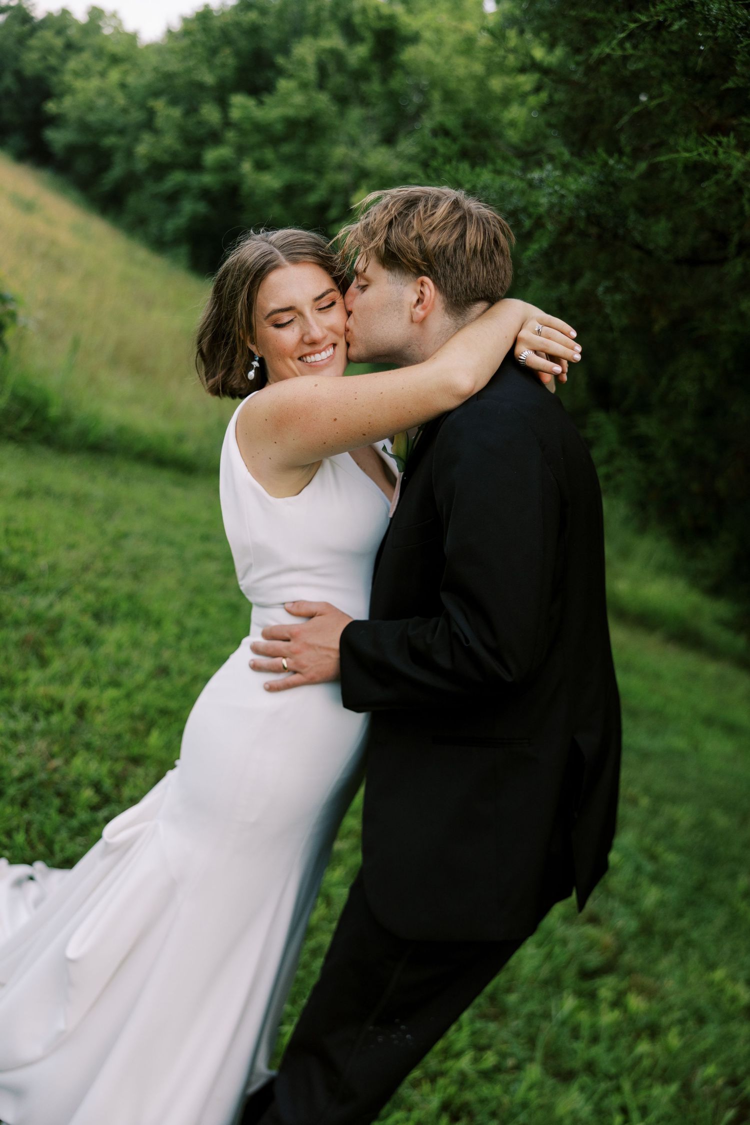 A couple shares a romantic moment in a lush green outdoor setting wearing formal wedding attire.