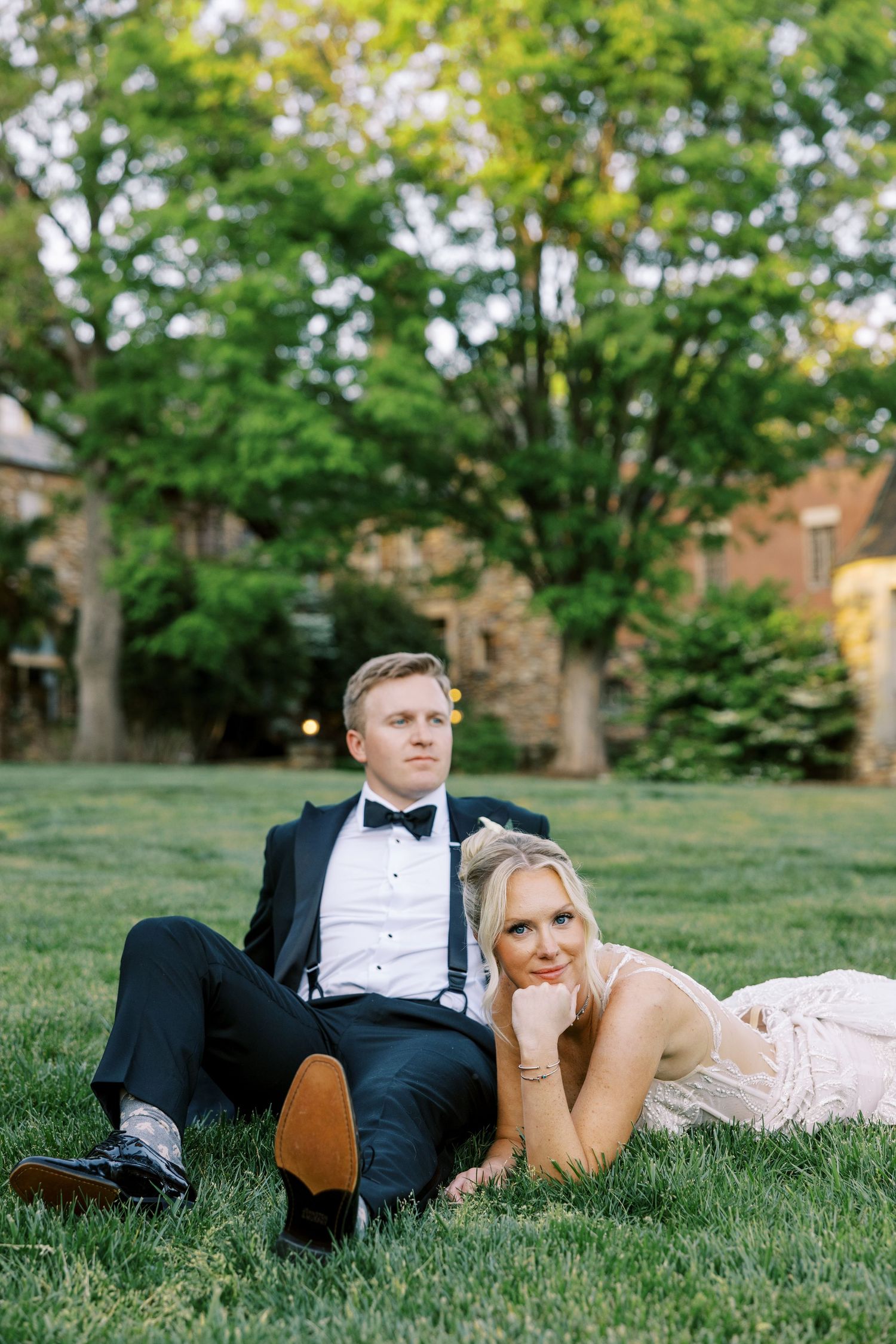 Bride and groom relaxing on green grass in formal wedding attire, surrounded by lush trees at golden hour.
