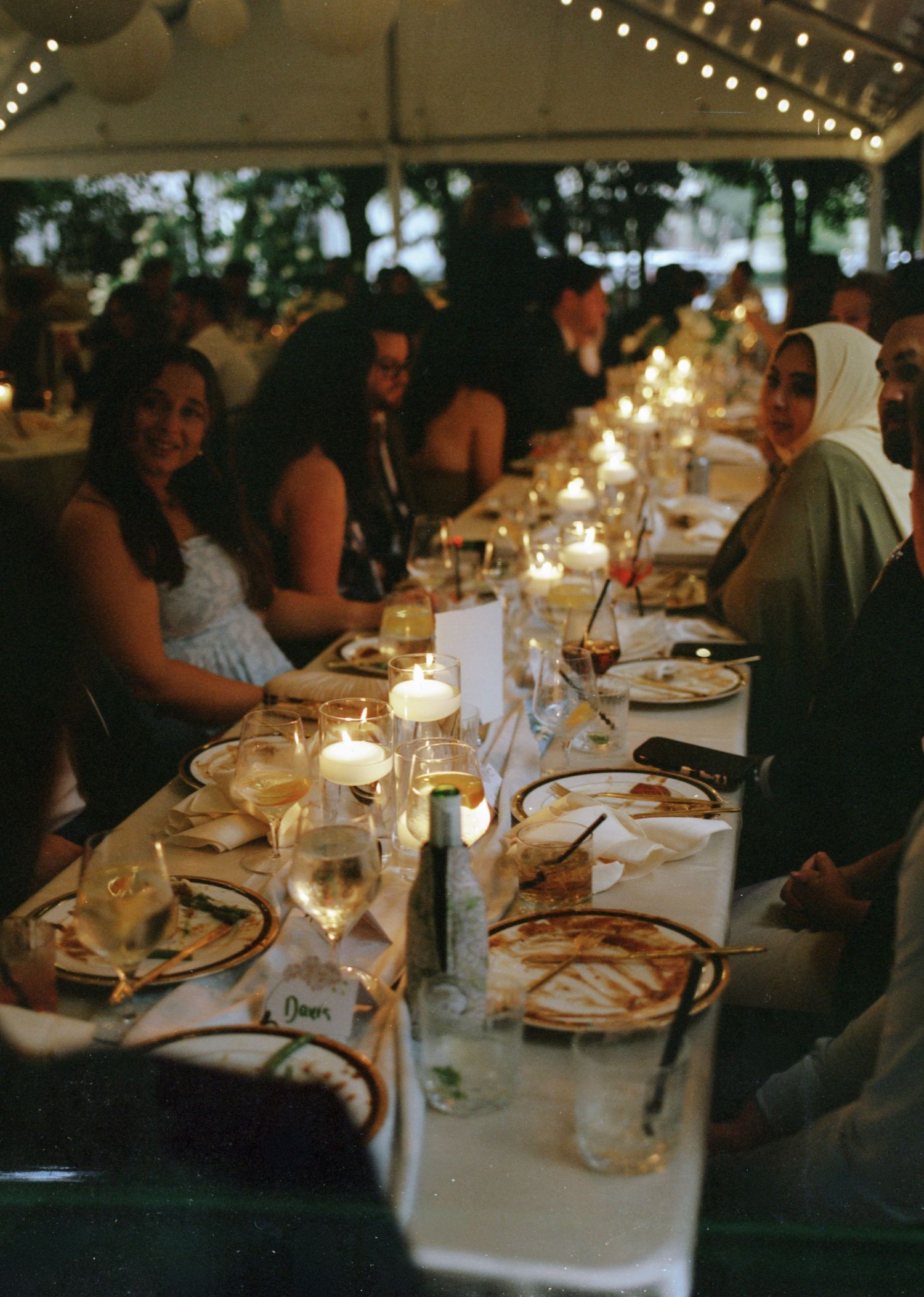 Guests seated at a long candlelit banquet table under a tent at an elegant outdoor evening wedding reception.
