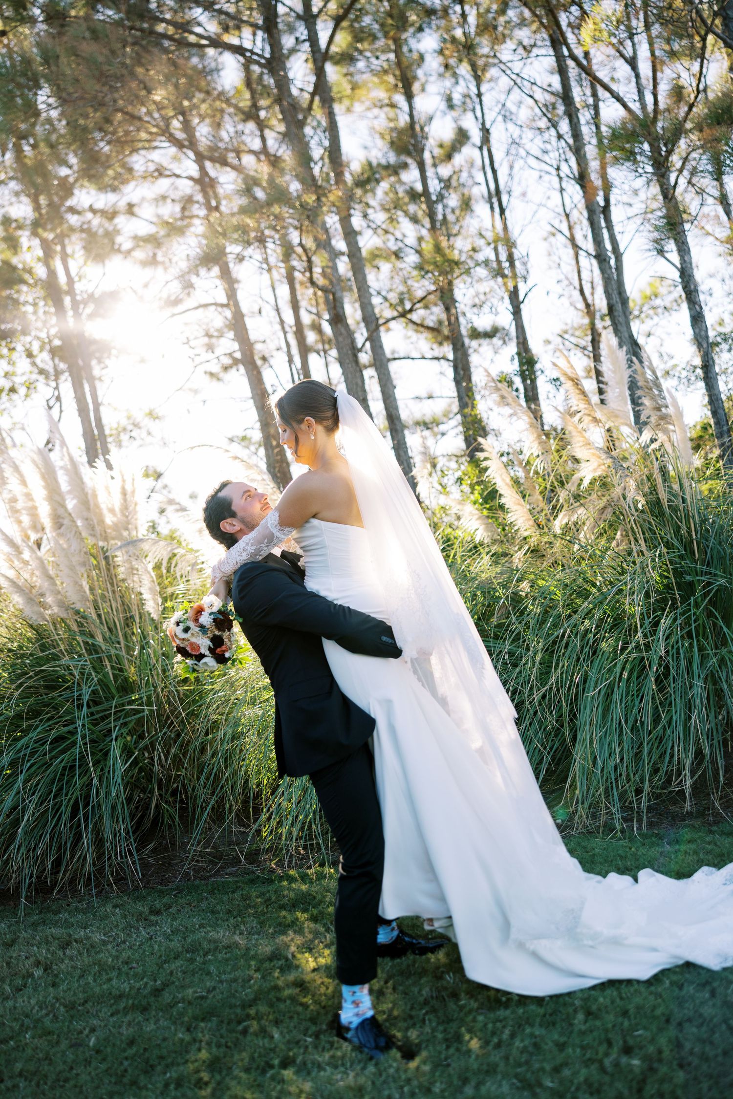 Romantic wedding couple embraces in a sun-drenched forest setting with tall grass and trees creating a magical backdrop.