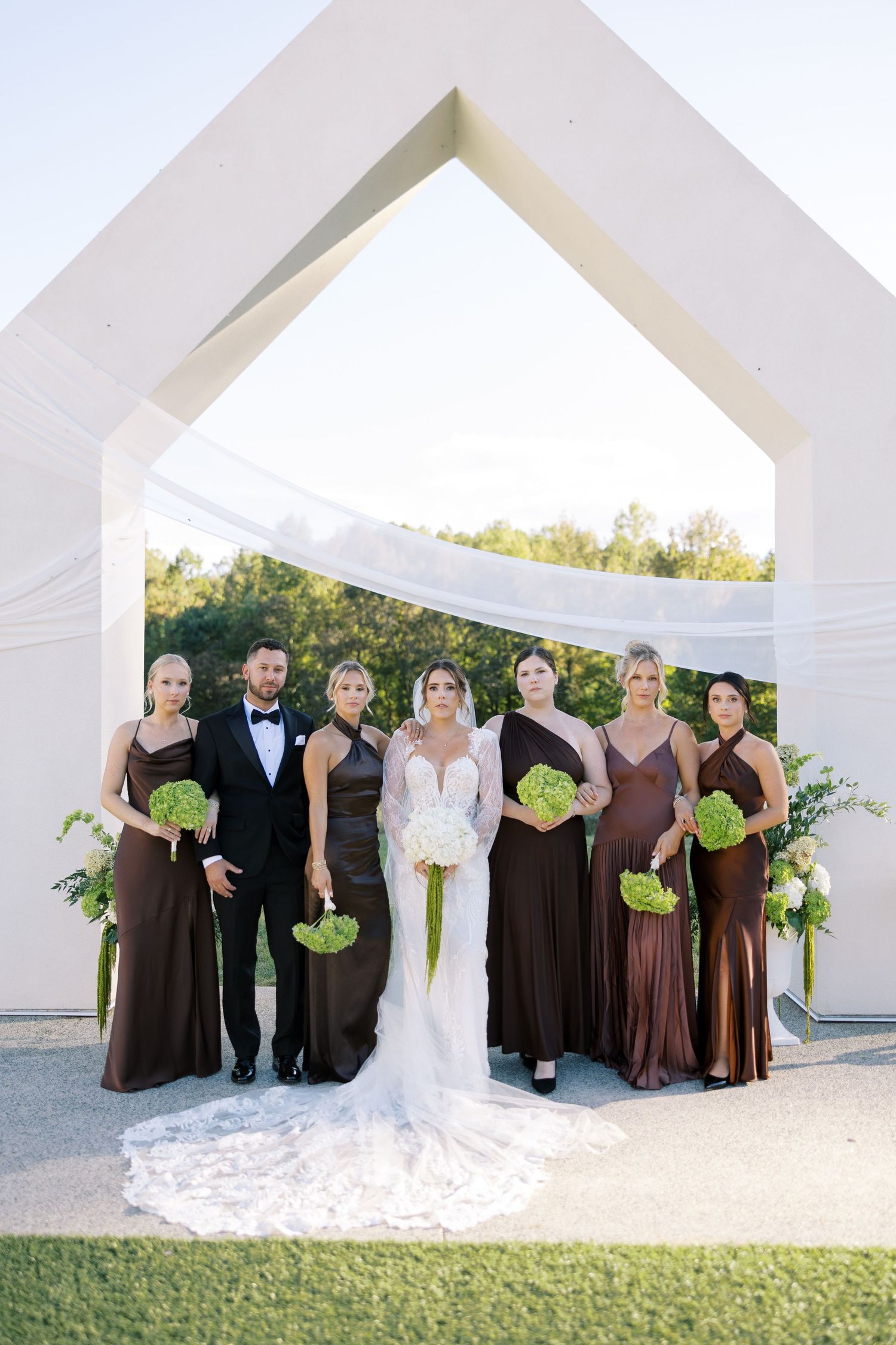 Wedding party poses in elegant brown dresses and formal attire under white architectural arches at an outdoor venue.