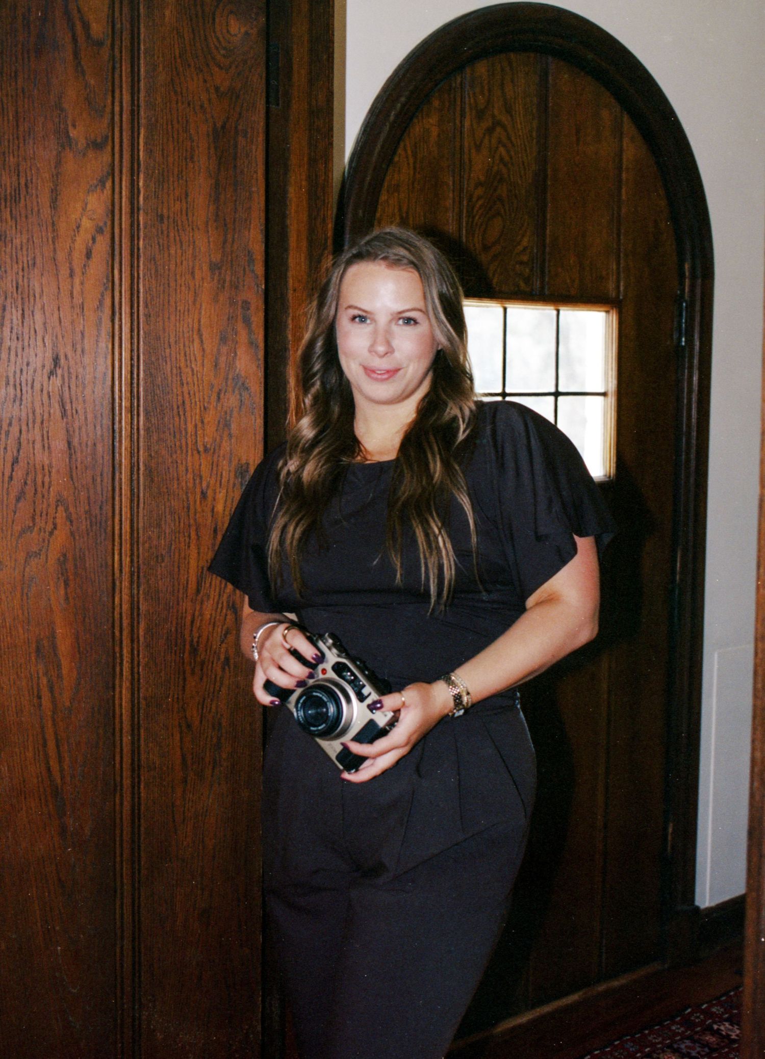 Woman in black outfit holding a camera stands in a wooden arched doorway, smiling at the camera.
