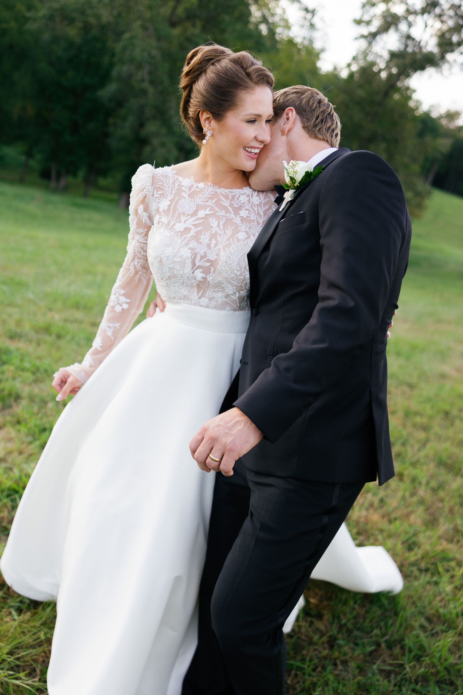 Bride in lace gown smiles as groom in black tuxedo whispers in her ear outdoors on green lawn.