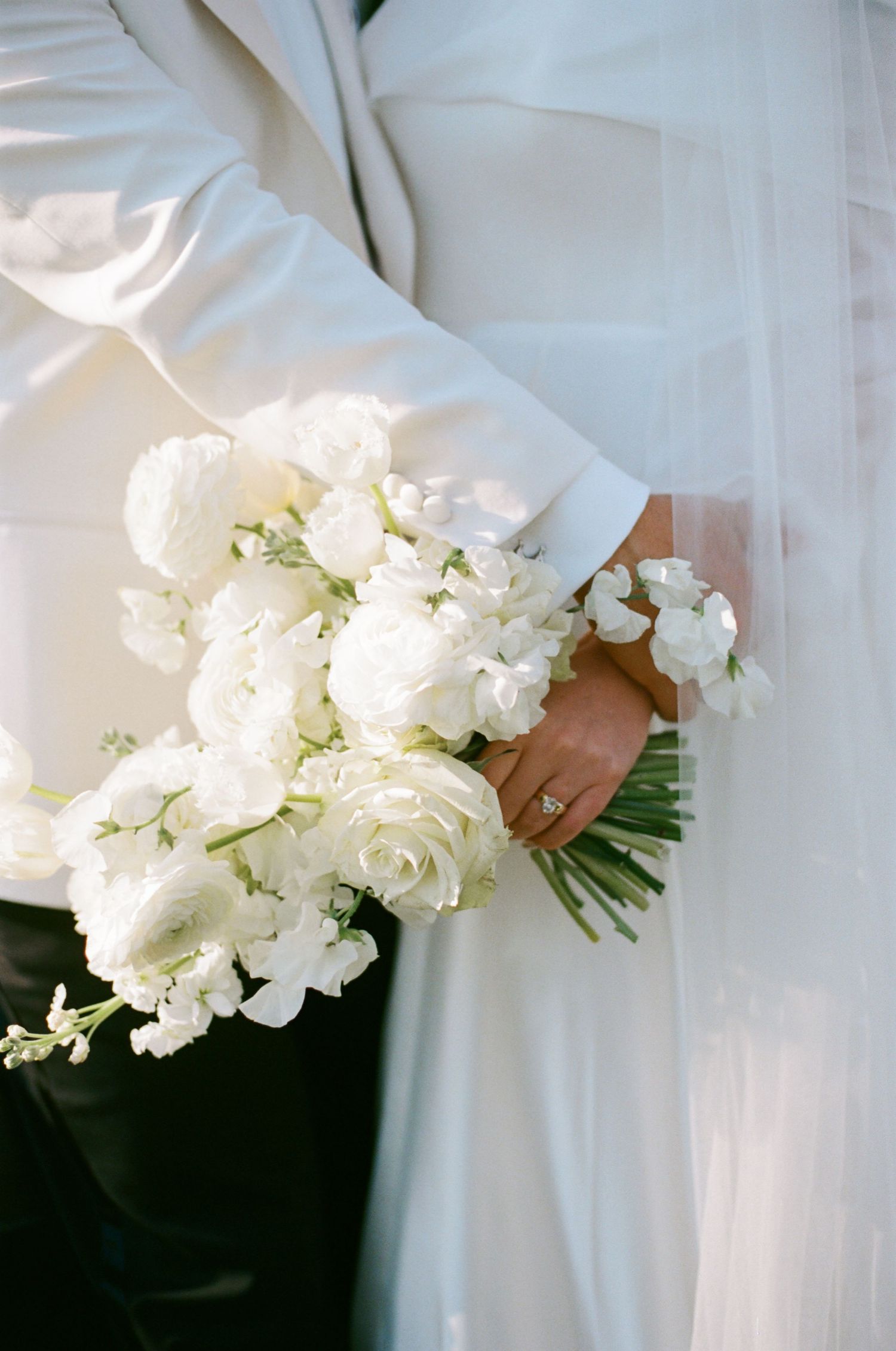 A delicate white wedding bouquet of roses and sweet peas held against a flowing white dress in soft natural light.