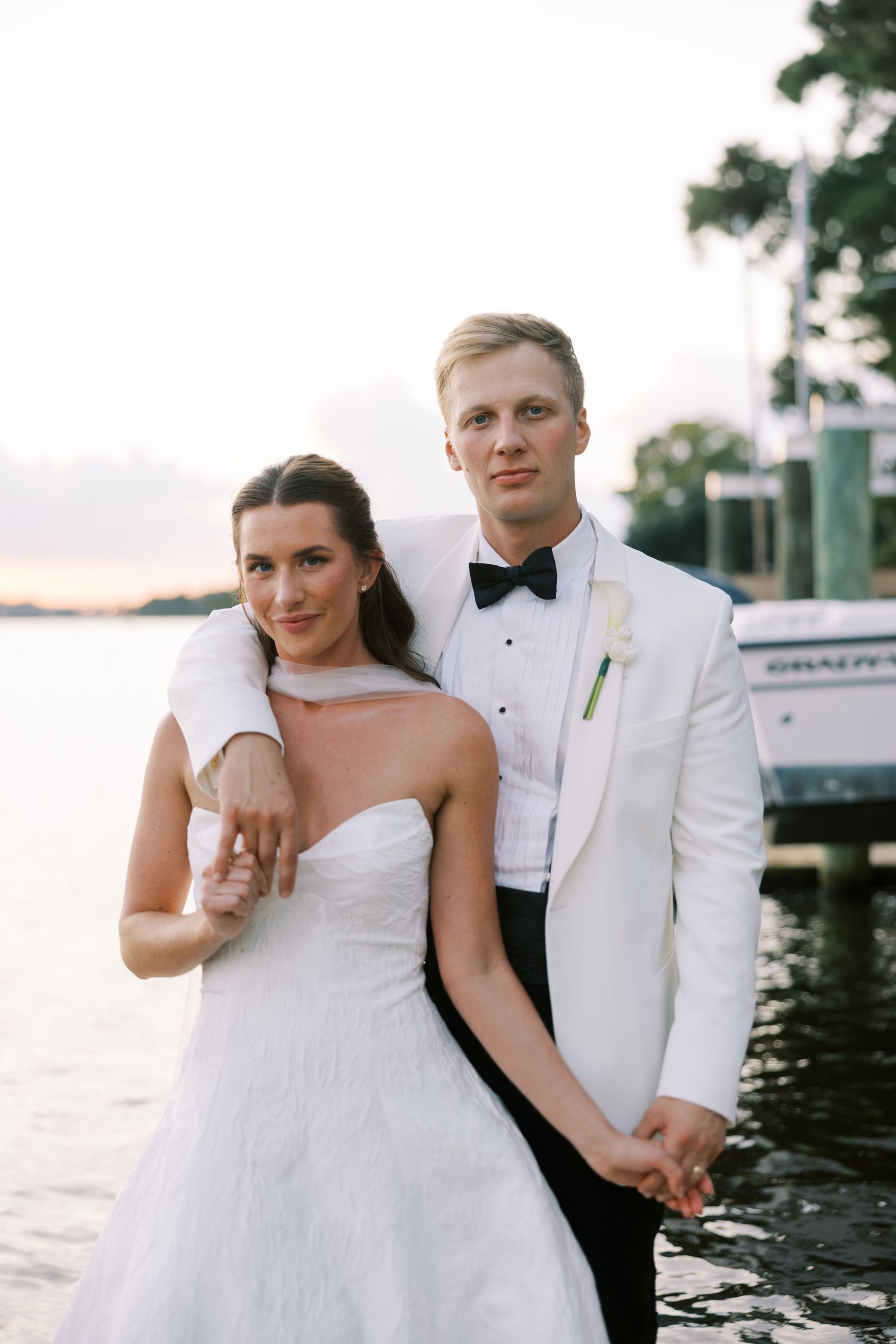 Bride and groom pose by waterfront at sunset, she in strapless gown, he in white tuxedo with black bow tie.