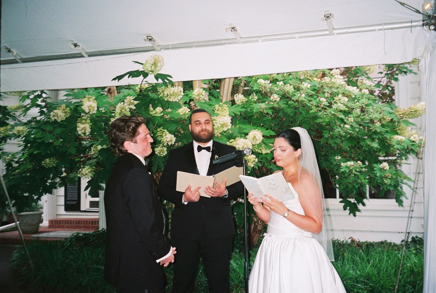 Bride and groom exchange vows at outdoor wedding ceremony under white tent with lush green floral backdrop.