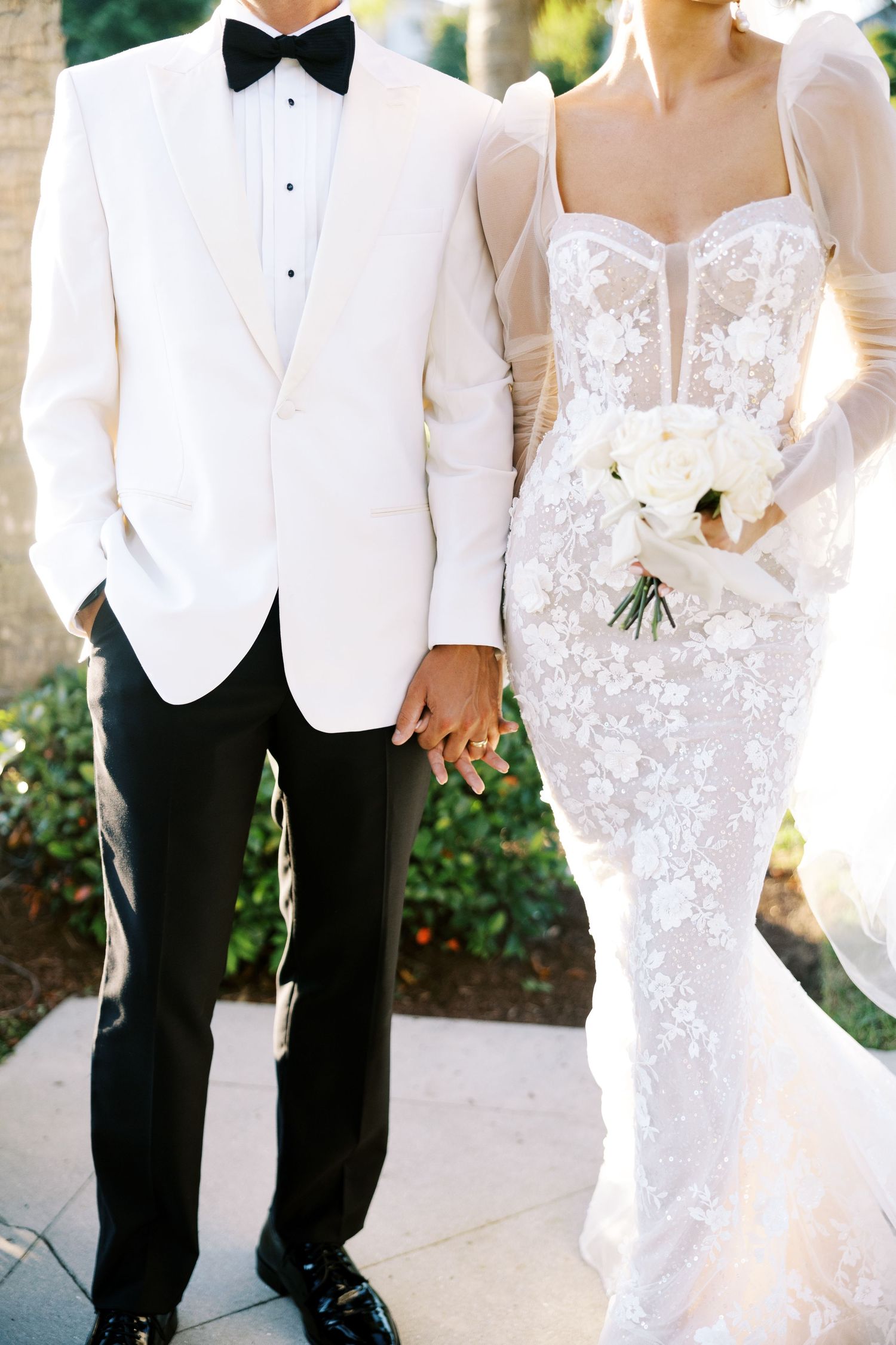 Bride in lace gown holding white bouquet stands beside groom in white tuxedo jacket and black bow tie.