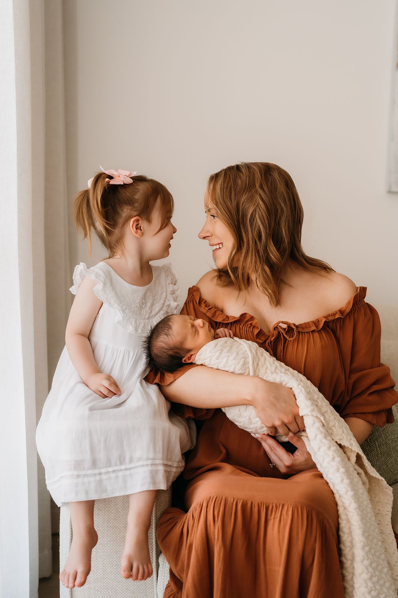 A smiling mother in a rust dress holds a newborn while her young daughter in white looks up at her lovingly.