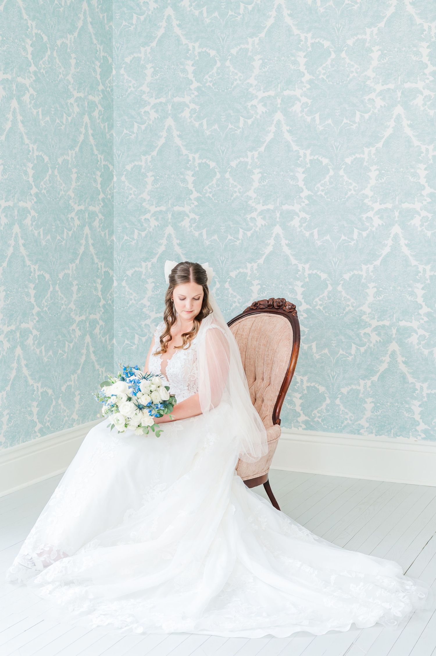 Bride sitting in a chair with blue wallpaper behind her looking down while holding a blue and white flower bouquet