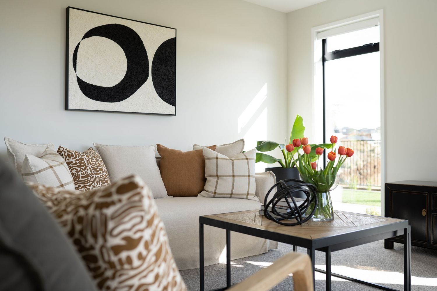 Modern living room with beige sofa, decorative pillows, abstract wall art, and red tulips on a wooden coffee table.