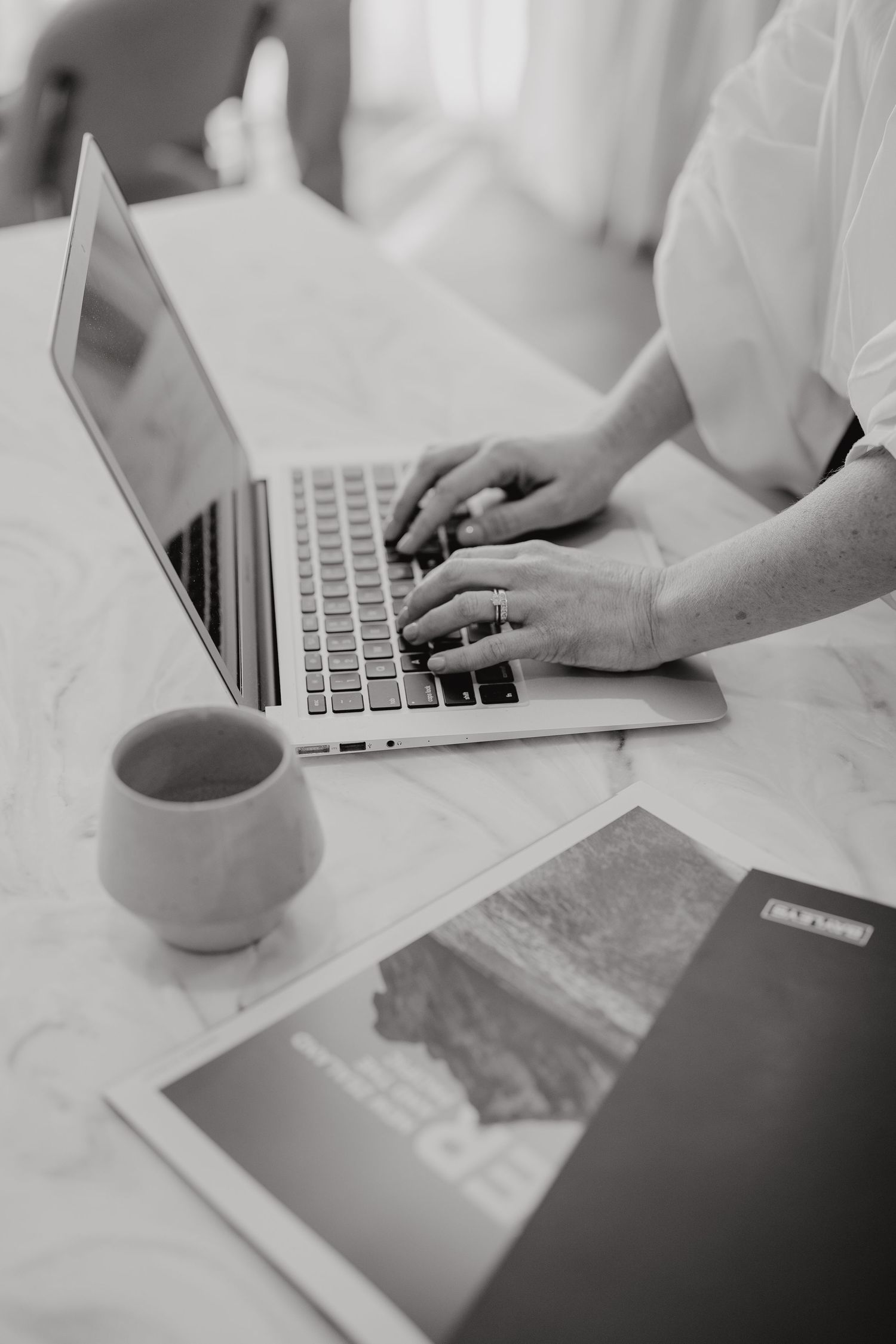 Person typing on laptop with coffee cup and tablet on white desk in black and white photography.