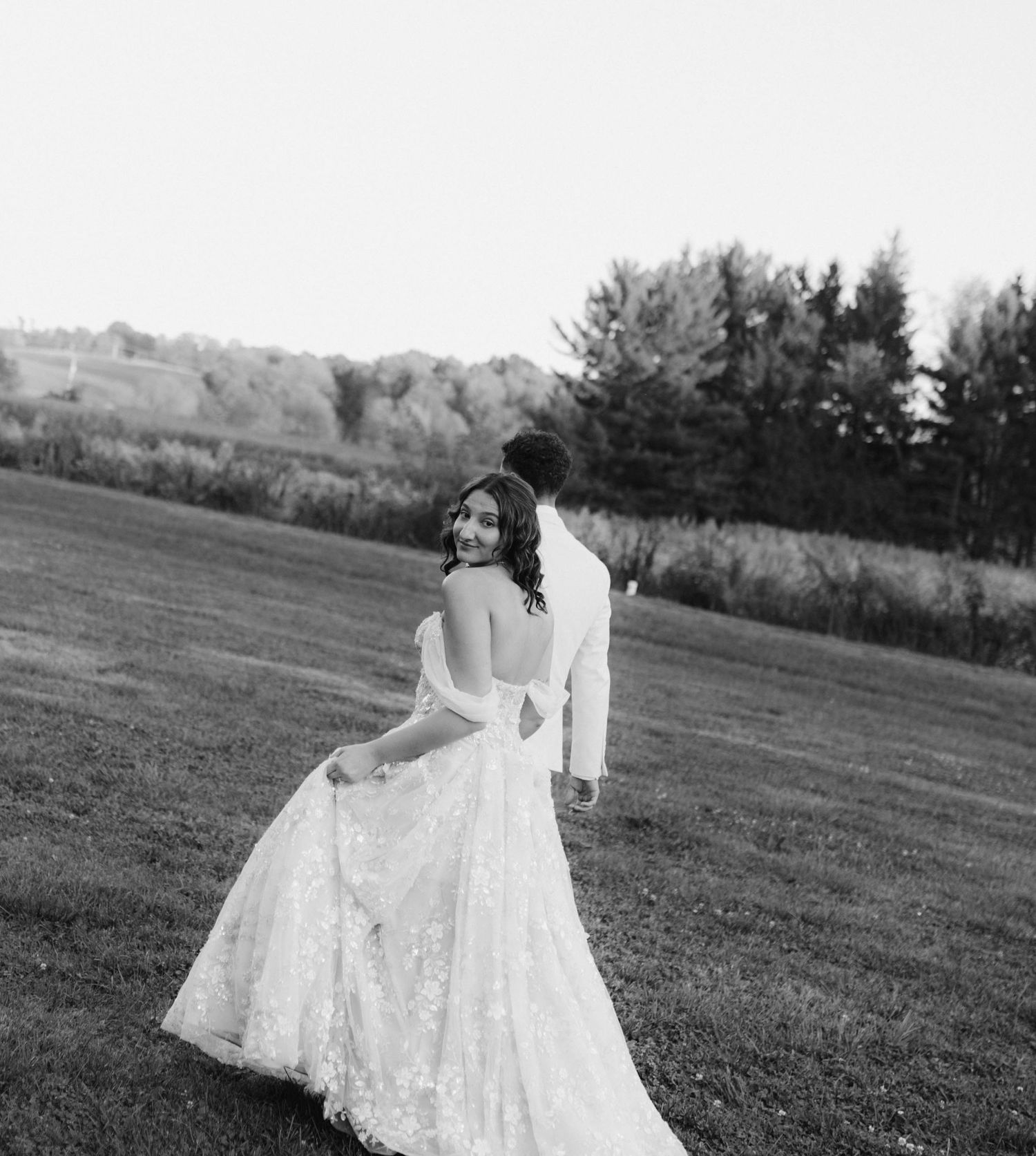 Black and white photo of bride in lace gown looking back at camera while walking with groom across green field.