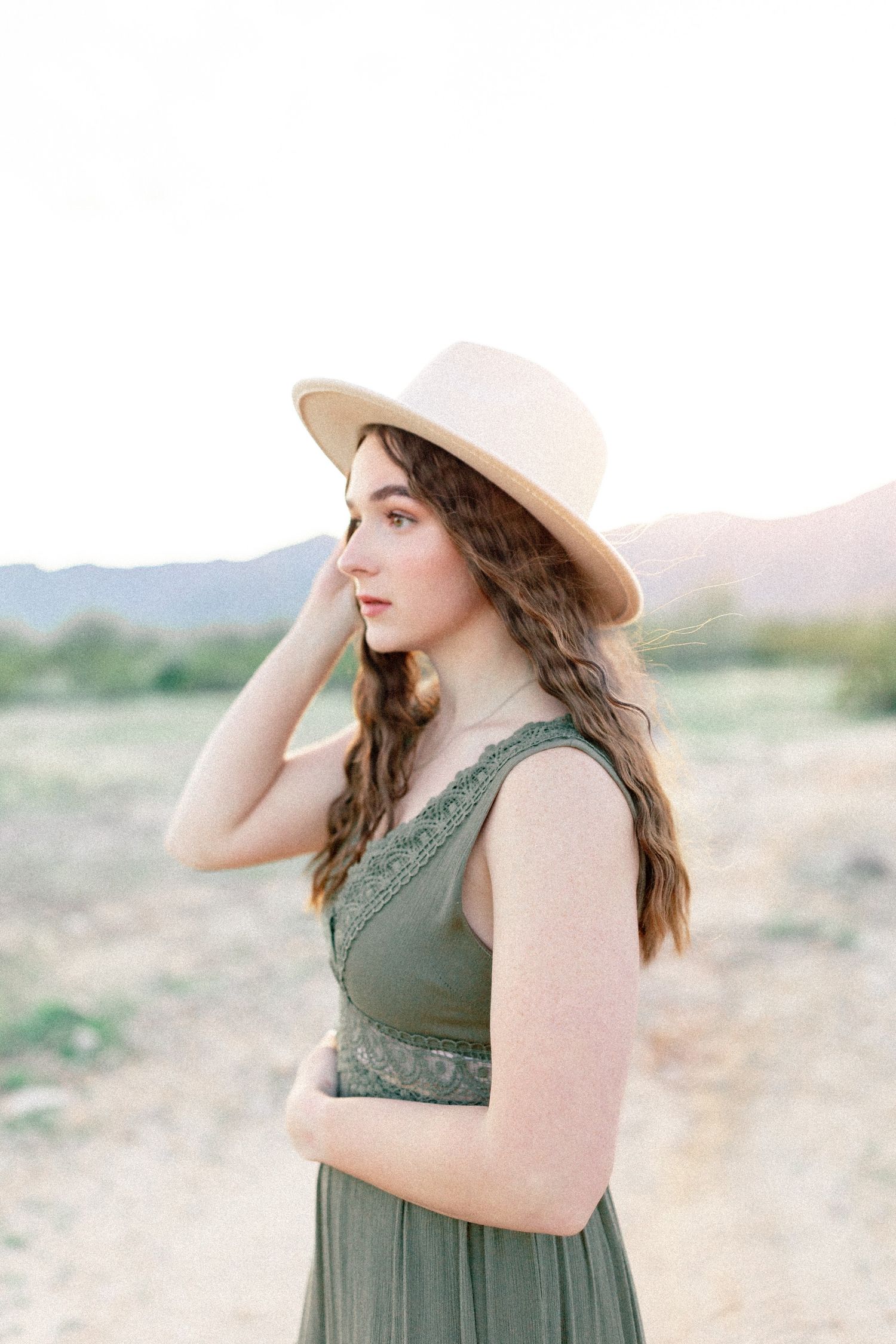 A person in a cream-colored hat and olive green dress stands in a desert landscape during golden hour.