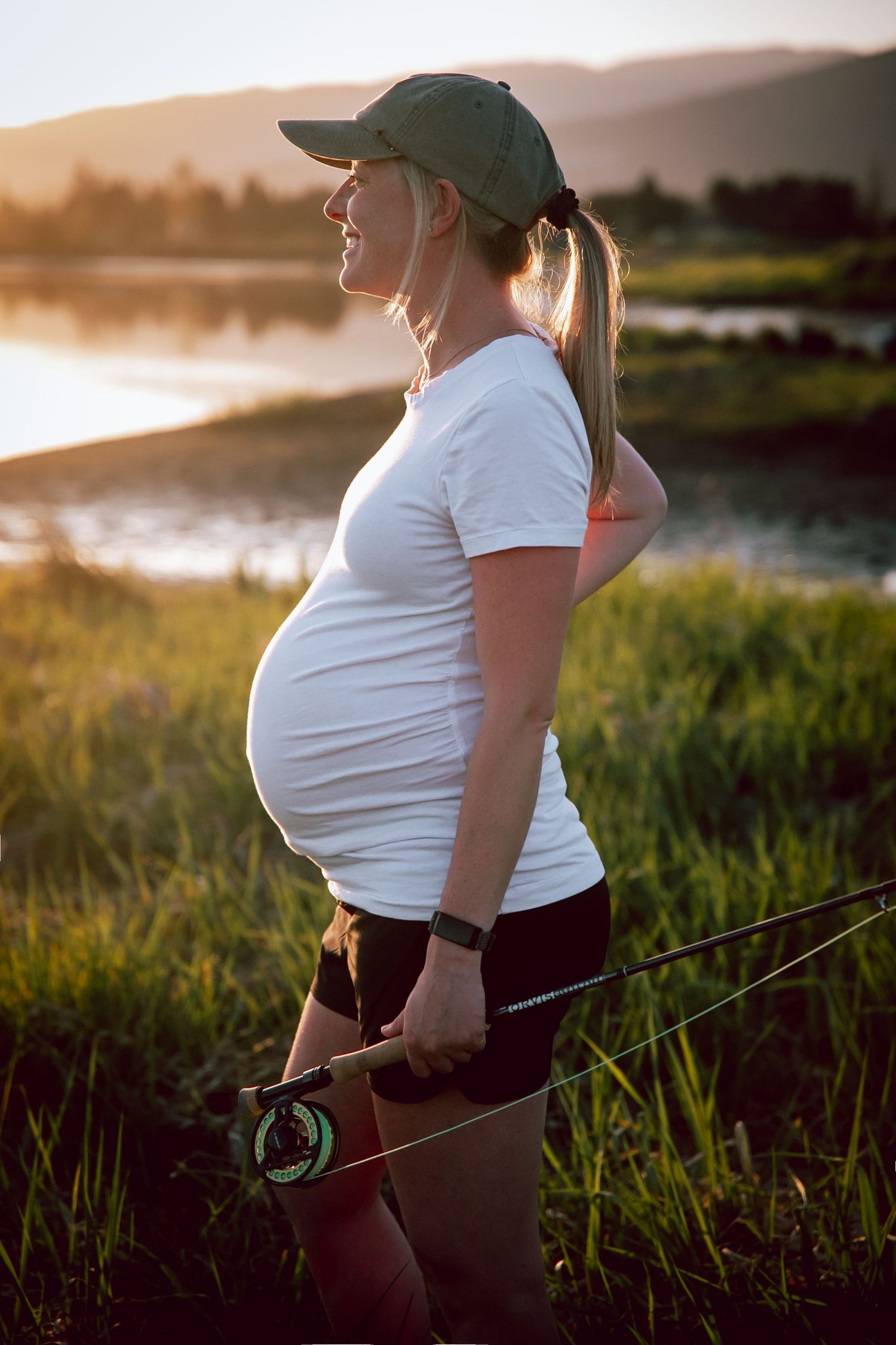 Pregnant angler in baseball cap holds fishing rod at sunset by scenic lake.