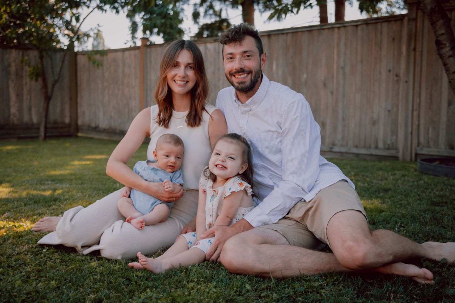 Family smiling together while sitting on grass in backyard during golden hour.