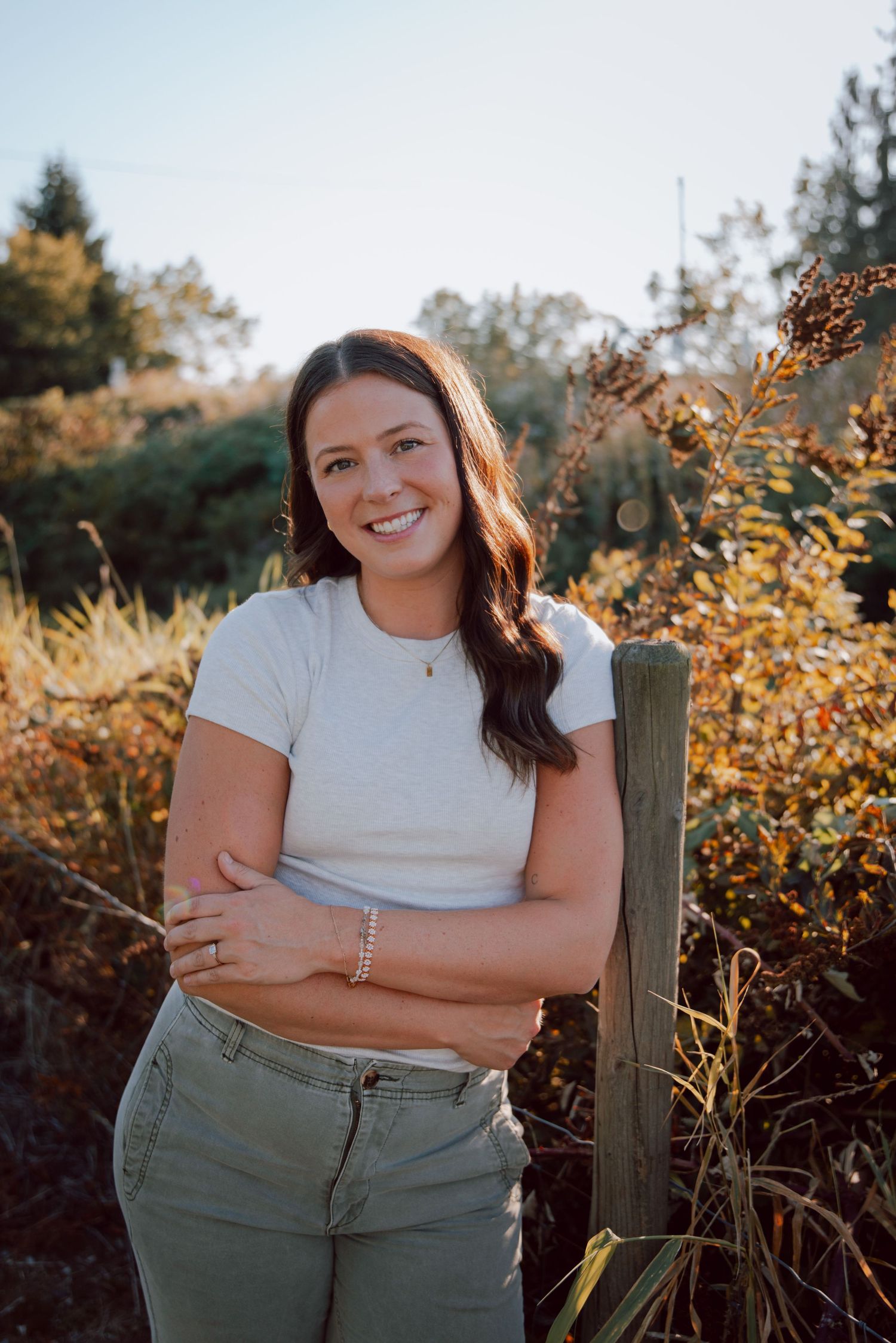 A person in a white t-shirt and olive pants poses against an autumn backdrop during golden hour.