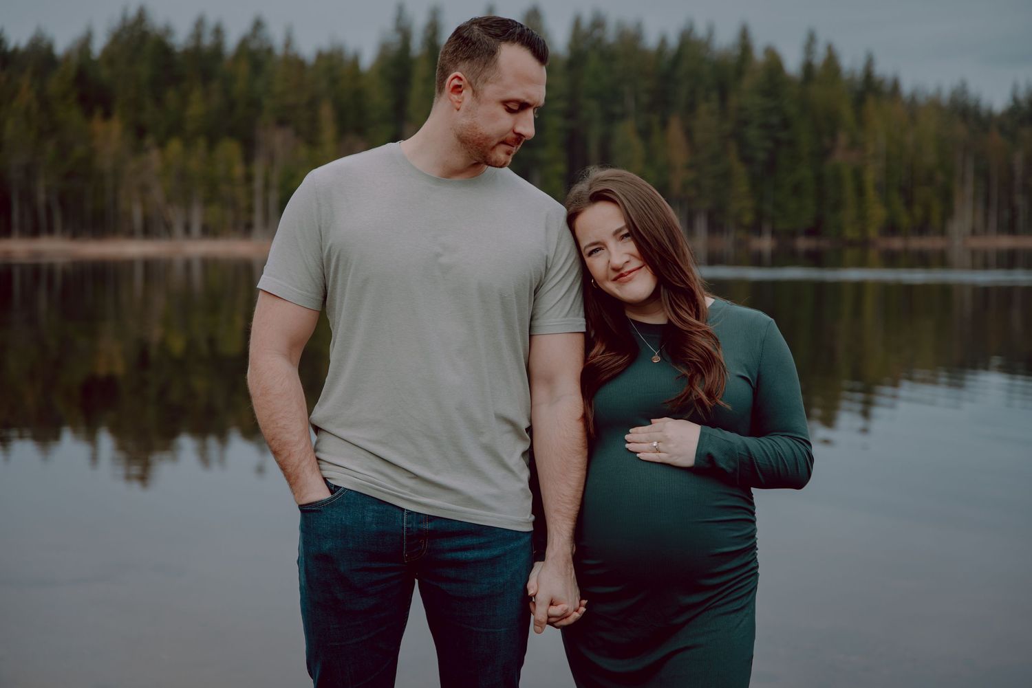 Expecting couple shares intimate moment by serene lake with evergreen forest backdrop.