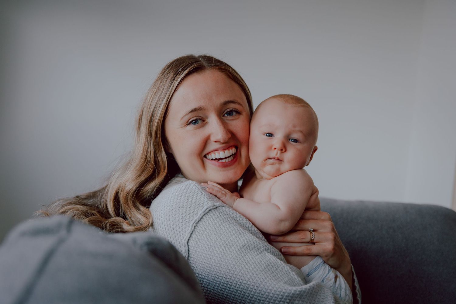 Parent and baby share a tender moment while relaxing together on a gray couch.