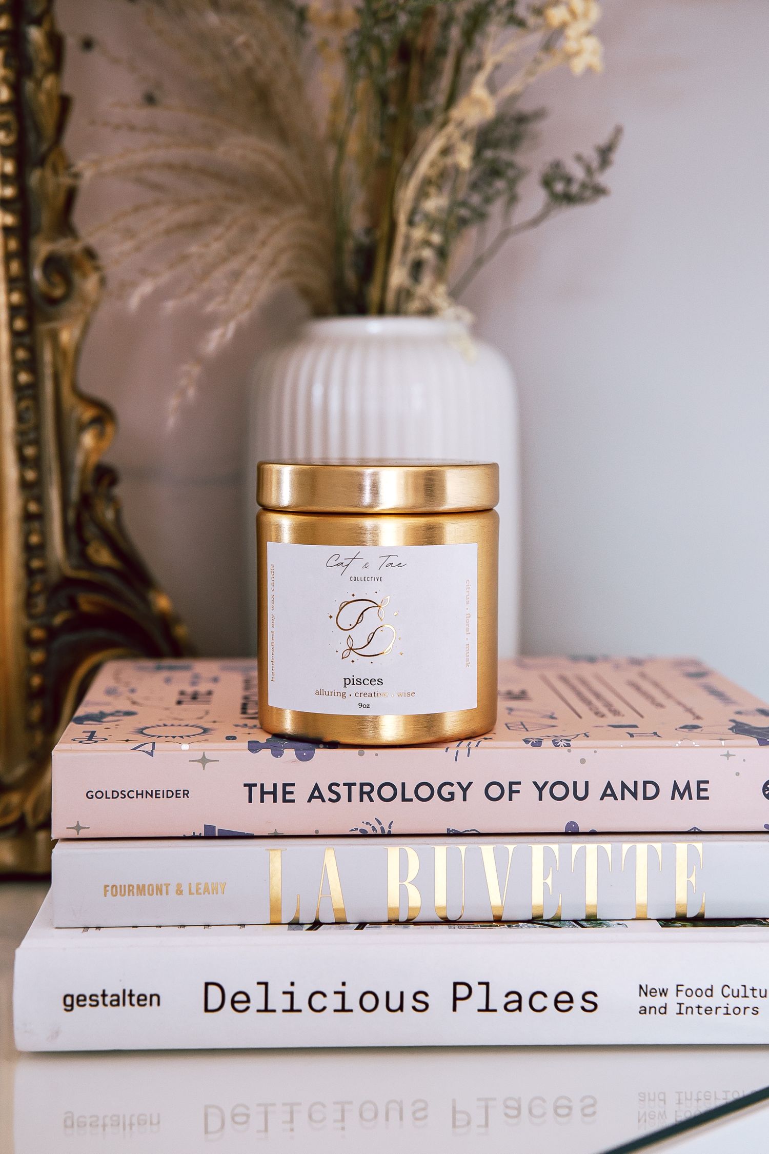 A styled product photo of books and candle jar with gold lid beside white vase with pampas grass against light background.
