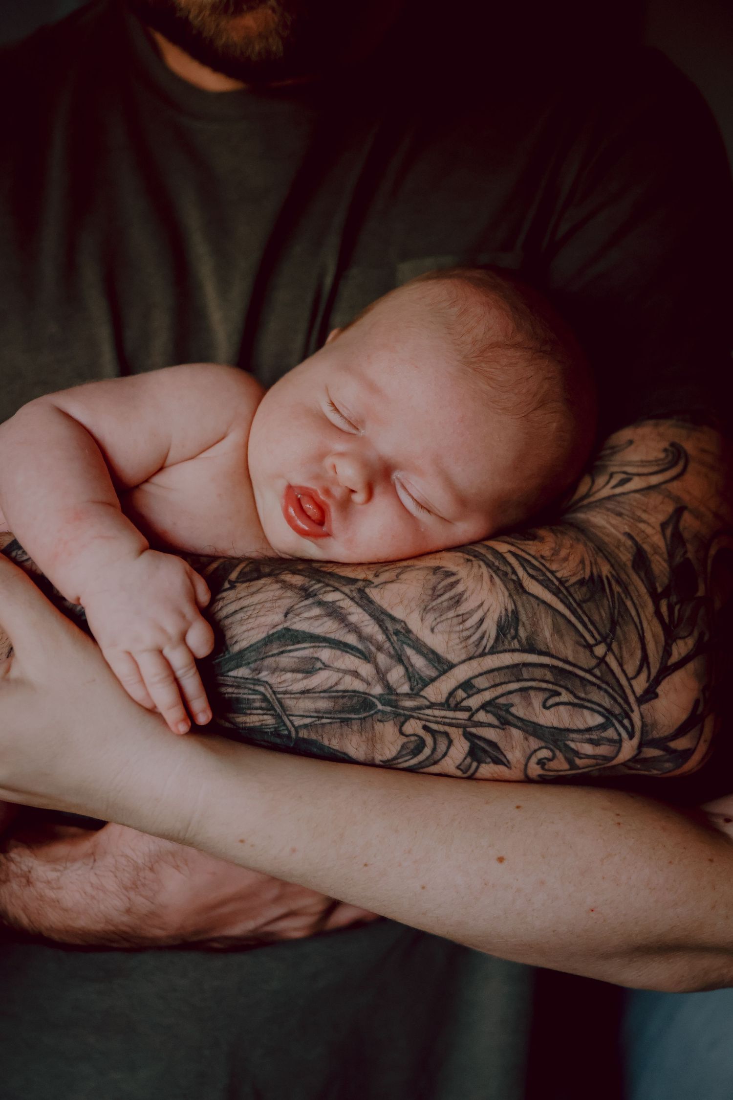 Sleeping newborn rests peacefully on tattooed arm in moody lighting.