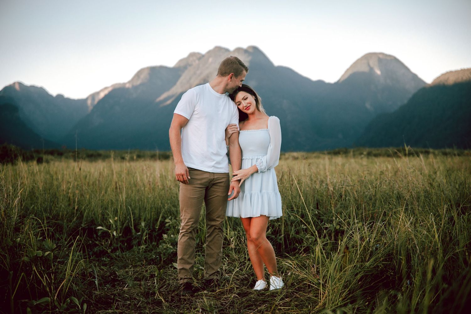 Couple standing together in grassy field with mountain views.
