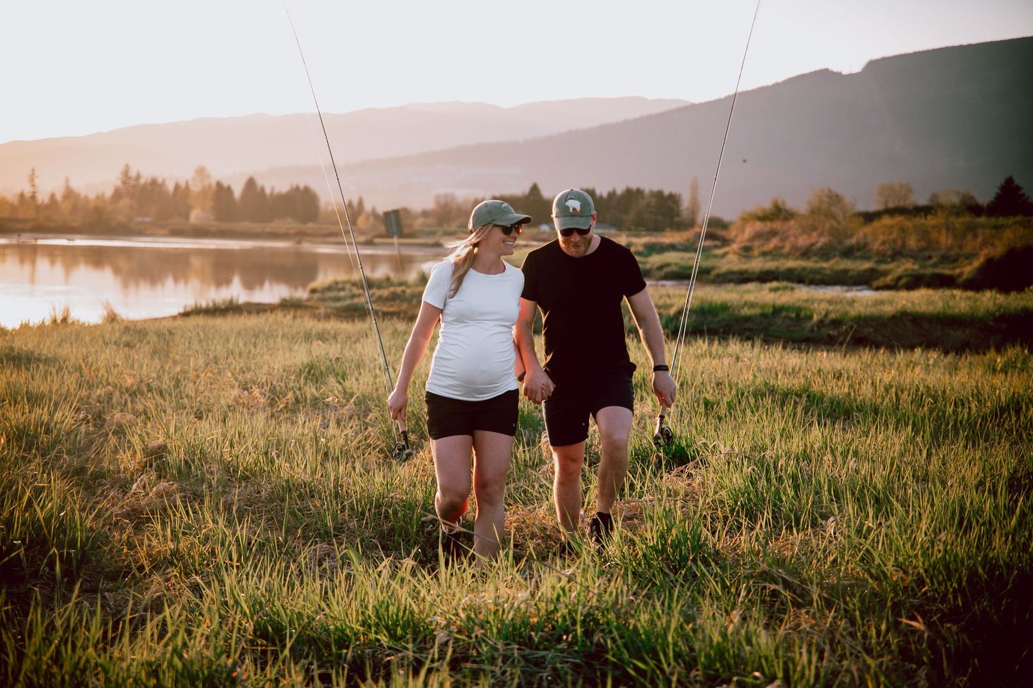 Couple walking together through tall grass at sunset near a peaceful lake.