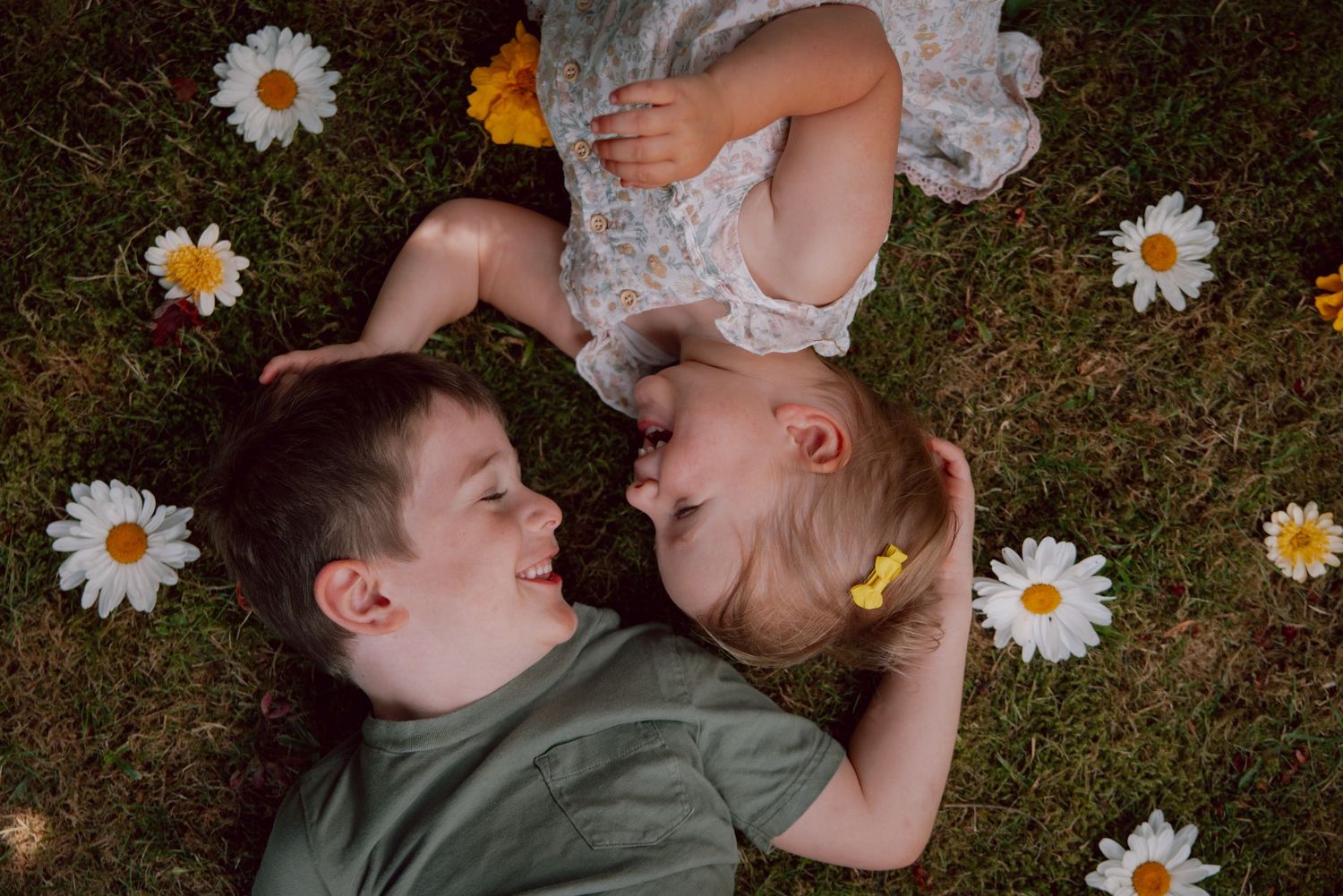 Couple lying on grass surrounded by scattered white daisy flowers in a romantic outdoor photoshoot.
