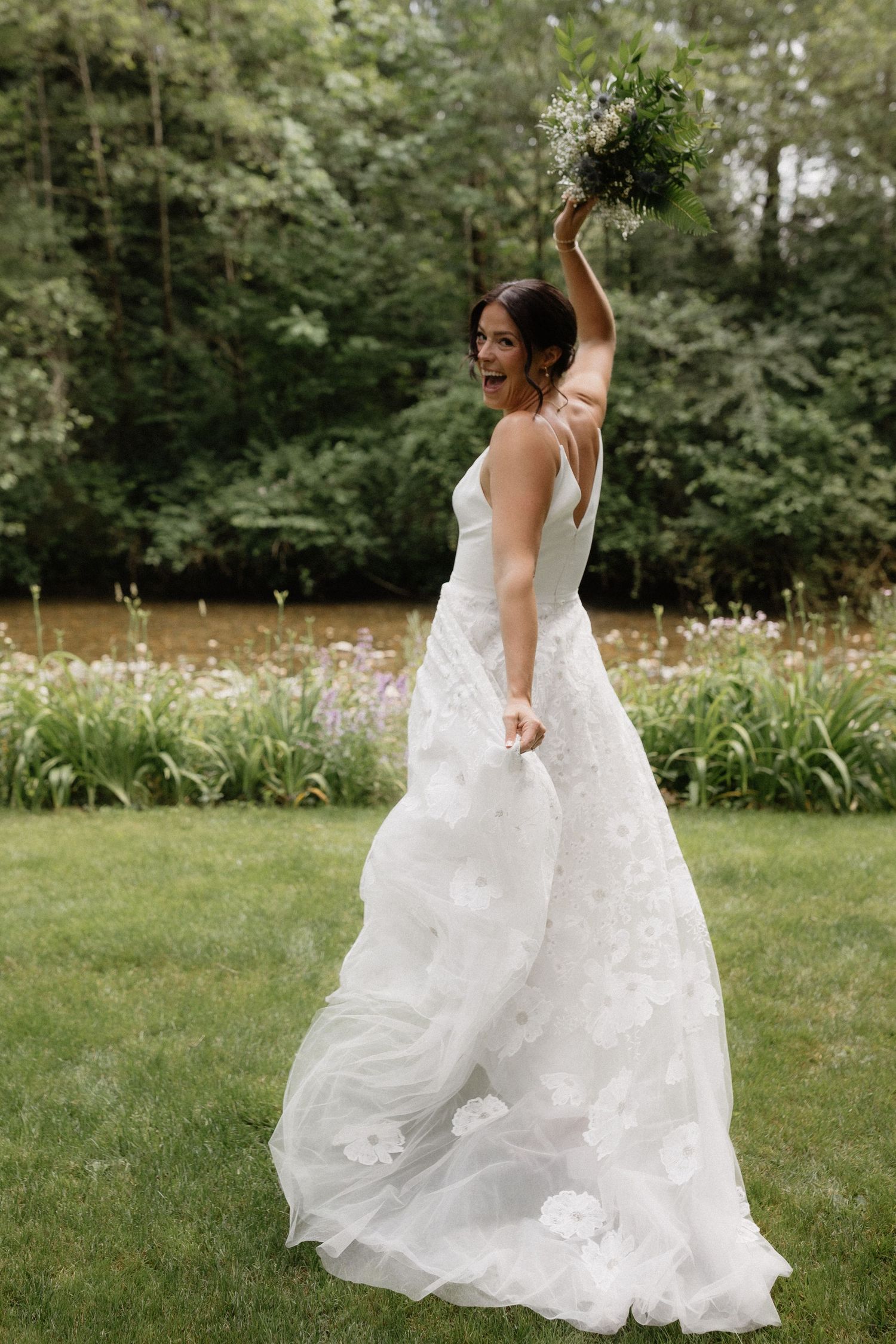 Bride in flowing white dress joyfully raises bouquet in garden setting.