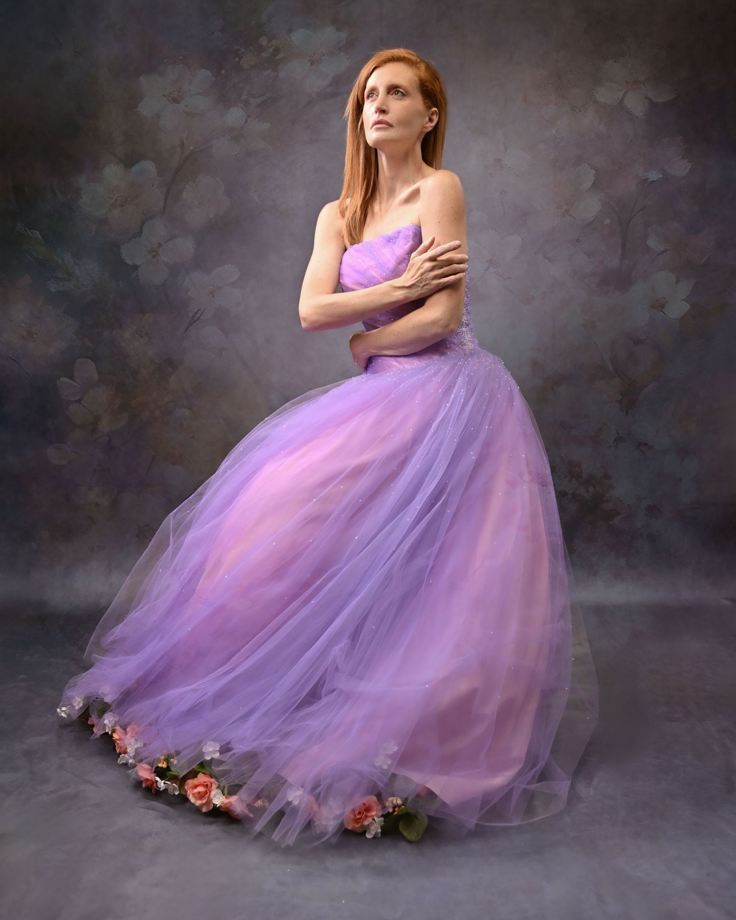 Dancer in lavender tulle gown poses dramatically against moody backdrop with flowers.