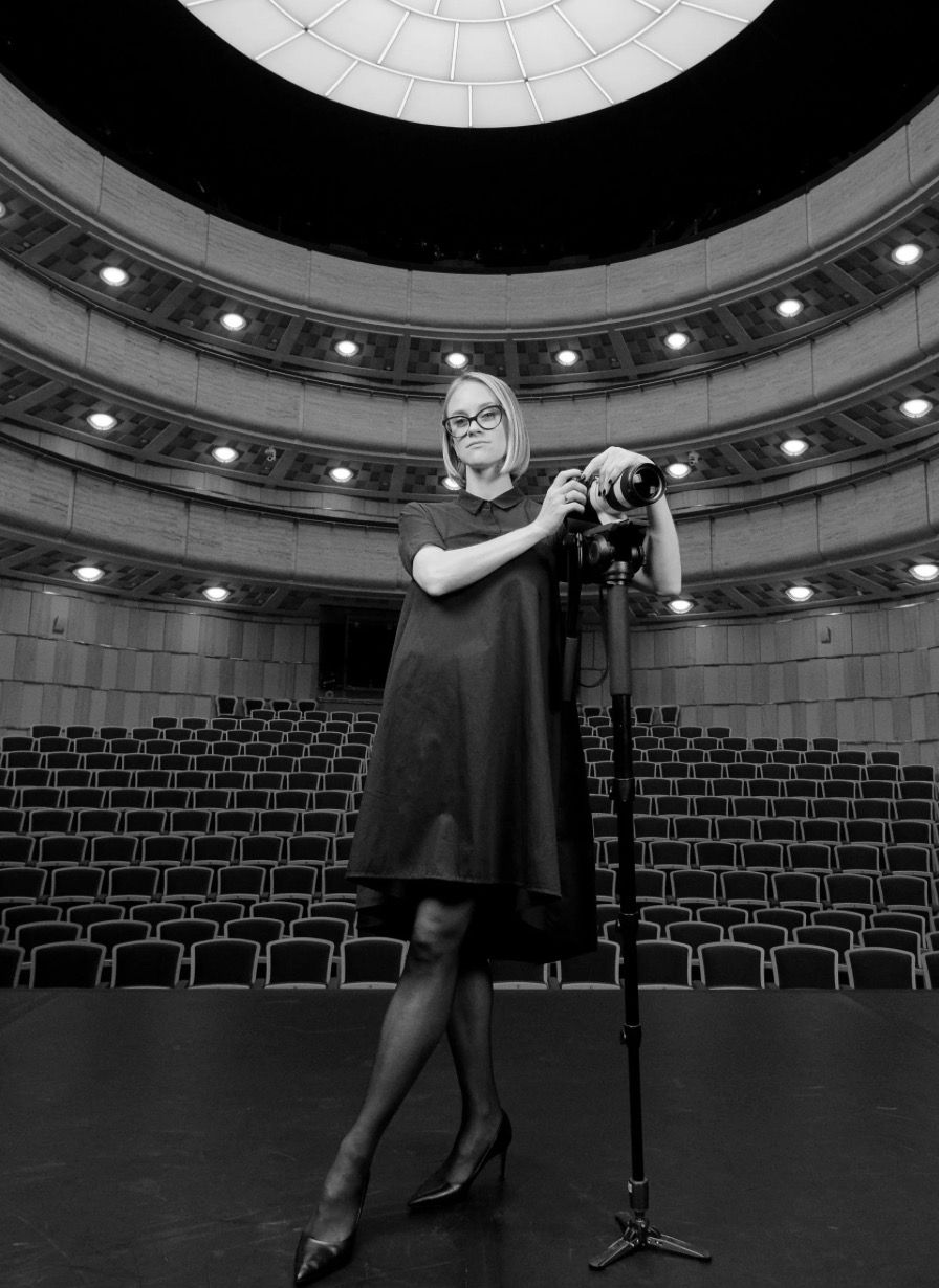 A photographer in a black dress stands with a tripod camera on an empty theater stage with curved seating rows behind.