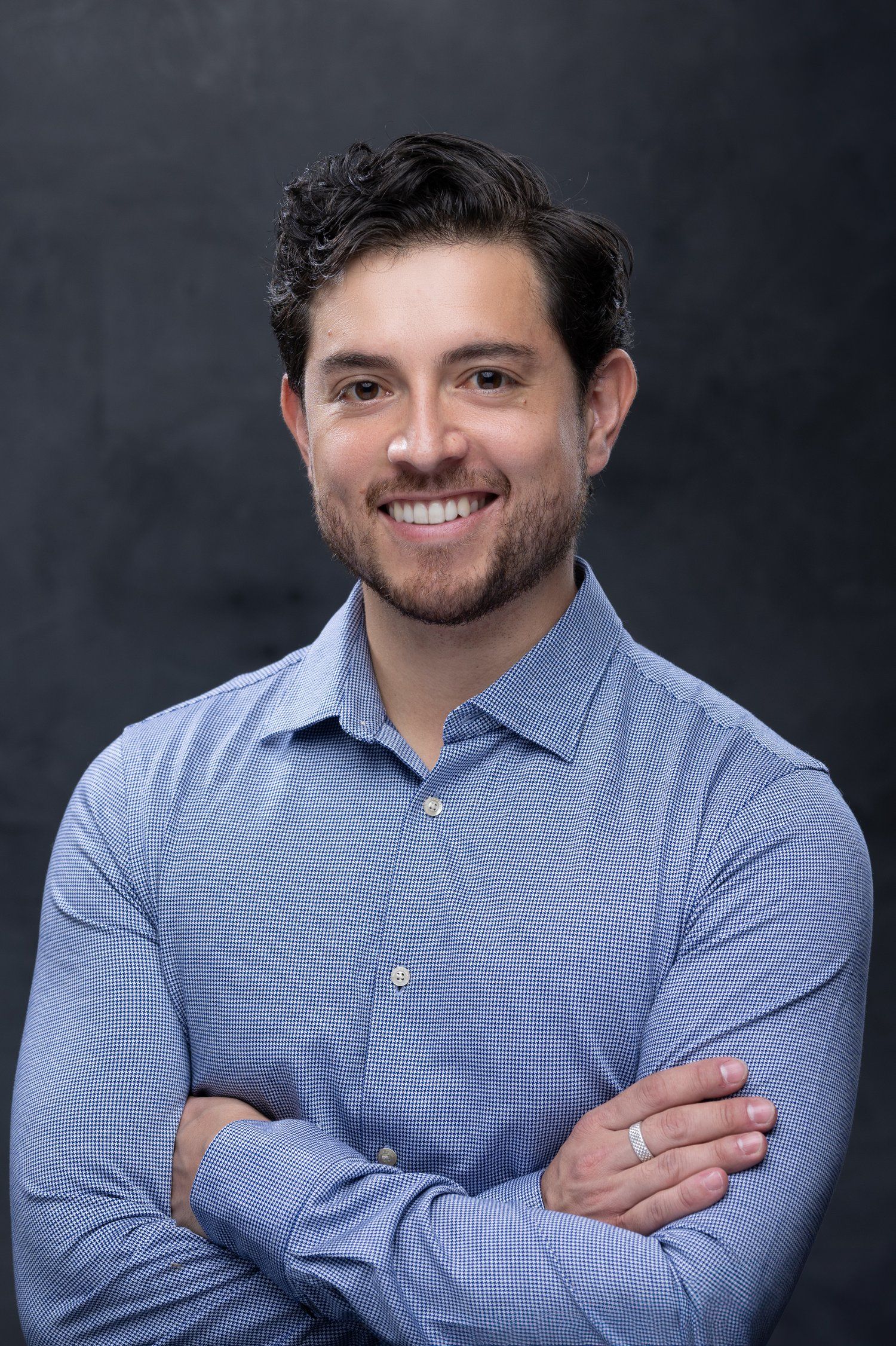 Professional headshot of someone wearing a light blue checkered dress shirt with arms crossed against dark background.