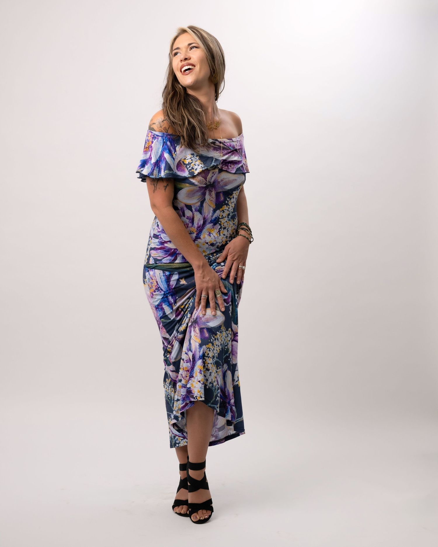 Woman in floral off-shoulder maxi dress with ruffles laughing against light background.