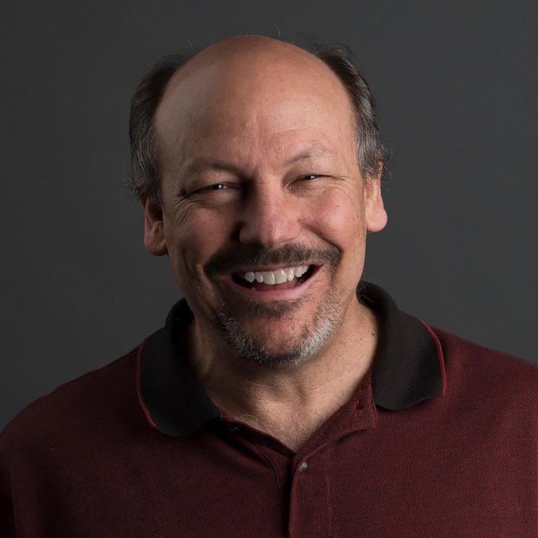 A person wearing a maroon polo shirt smiles warmly at the camera against a dark background.